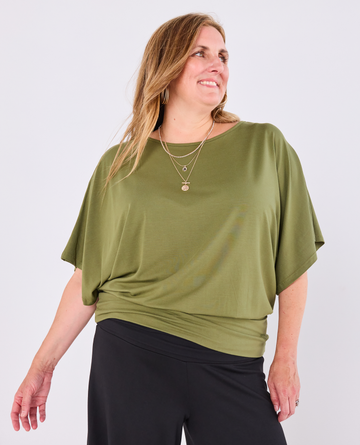 A woman wearing a Melyr, short-sleeve olive green Nico top and black pants, standing against a light grey studio background.