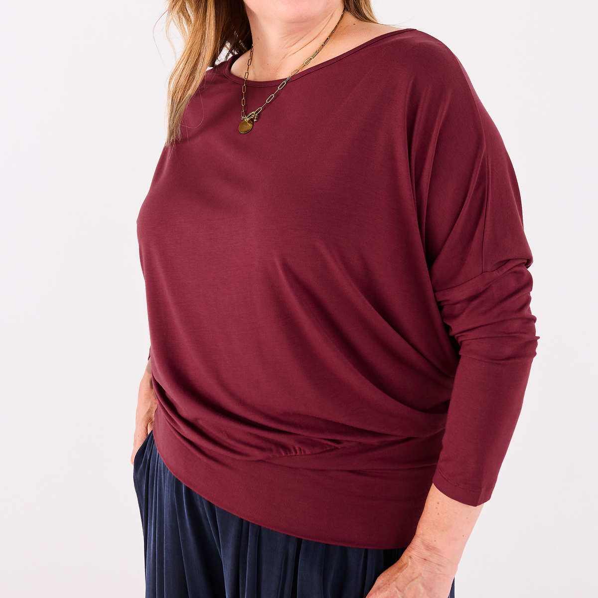 A woman wearing a long-sleeve, Melyr Aimee boat-neck jersey top in deep burgundy, paired with dark navy pleated pants, photographed against a clean white studio background.