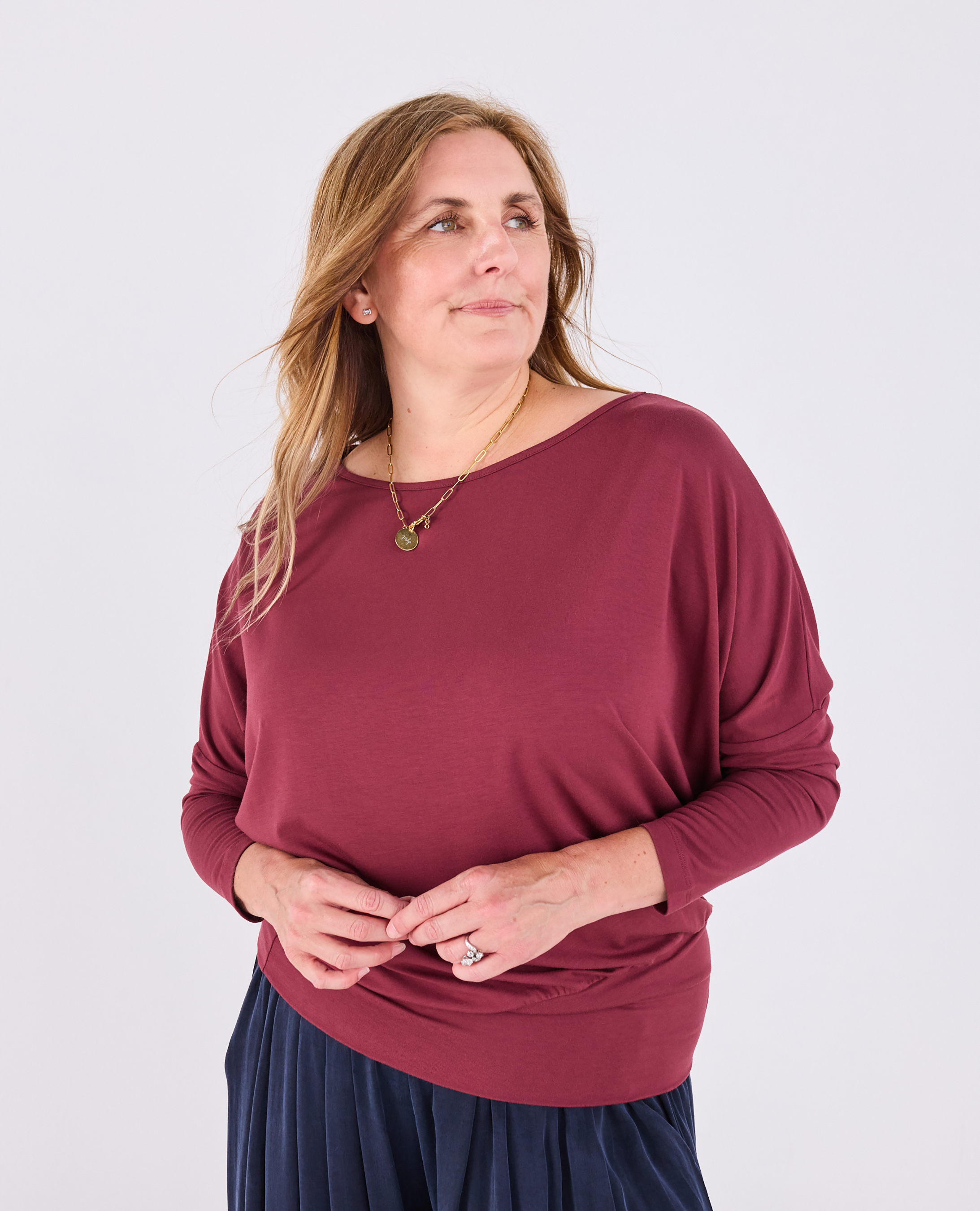 A woman modeling a long-sleeve boat-neck top in burgundy, paired with a navy skirt, against a light gray studio background.