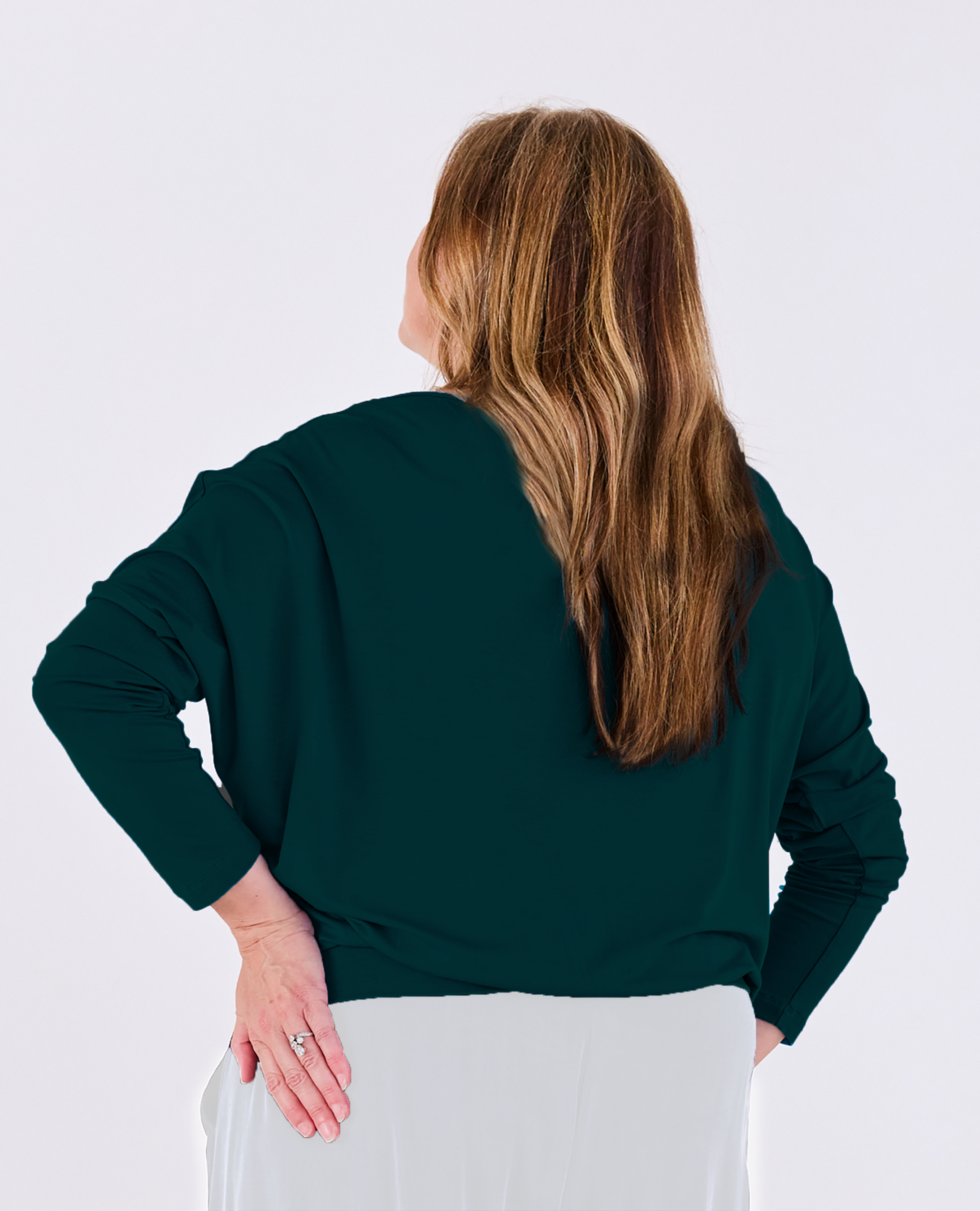 A woman viewed from the back wearing a dark green Melyr Aimee long-sleeve batwing top and white pants against a plain light grey studio background.
