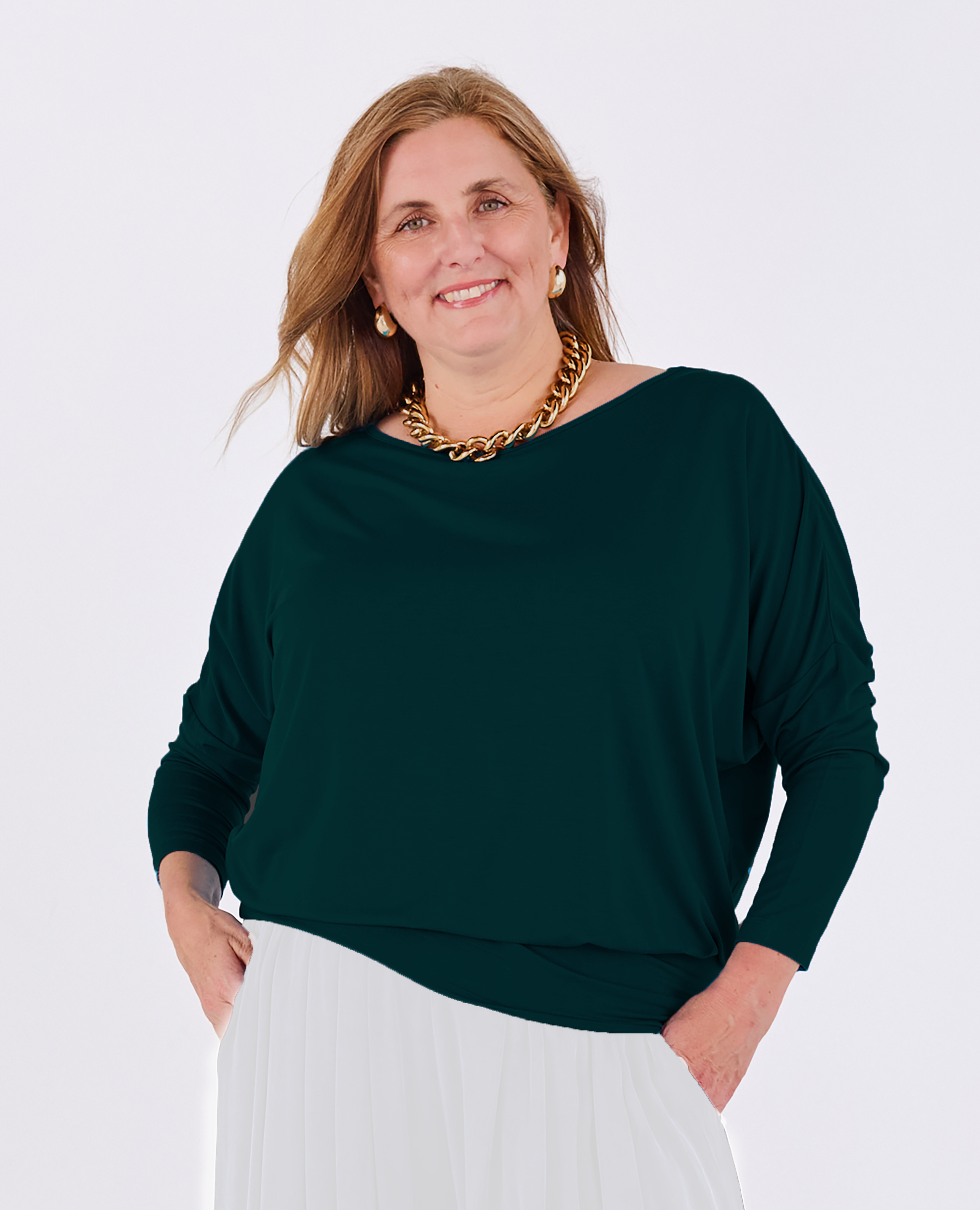 A woman wearing a dark green Melyr Aimee long-sleeve boat-neck top and white pleated skirt, accessorized with a chunky gold chain necklace and gold hoop earrings, posing against a light grey studio background.
