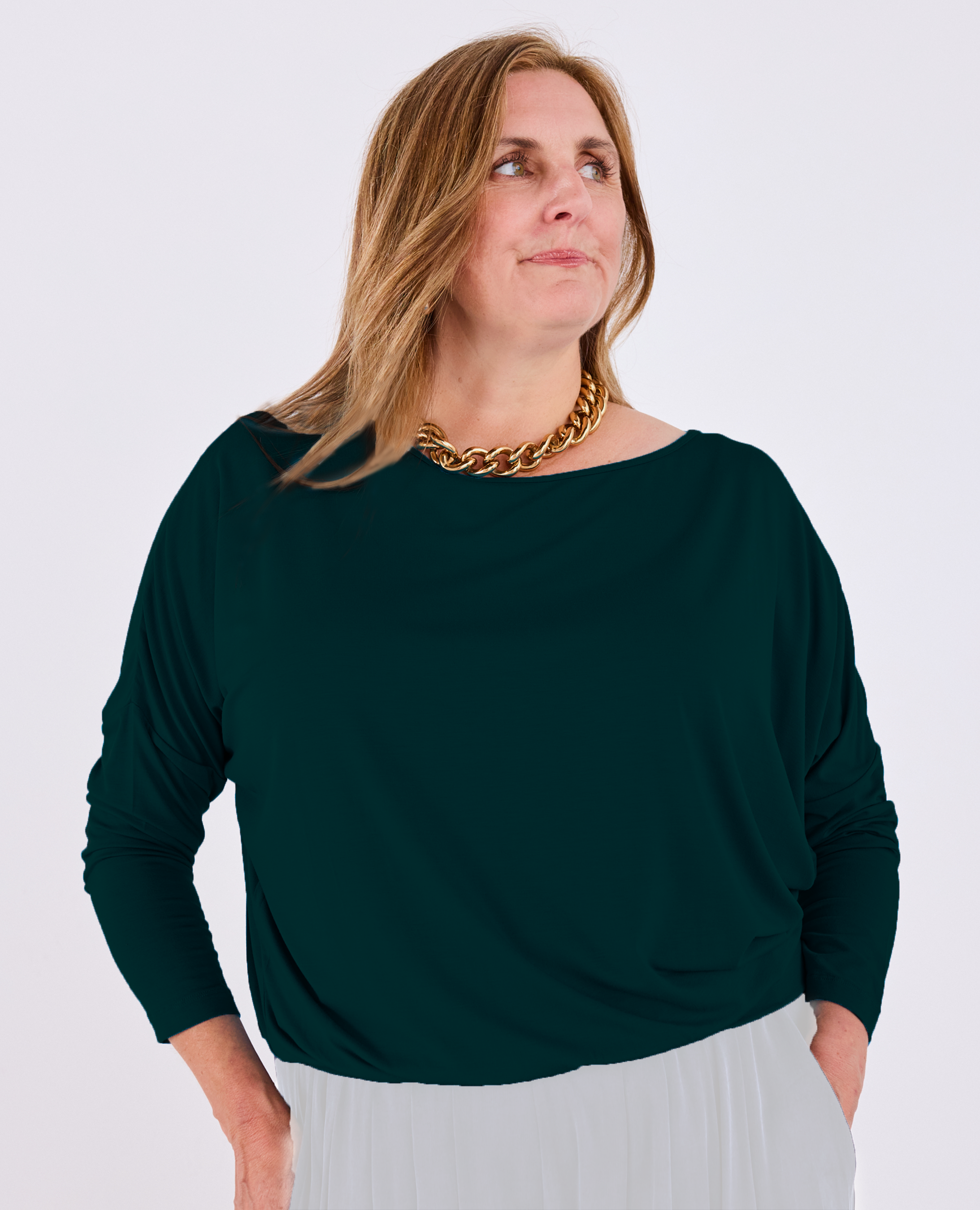 A woman wearing a dark green Melyr Aimee long-sleeve batwing top and a chunky gold chain necklace, photographed against a light grey studio background.