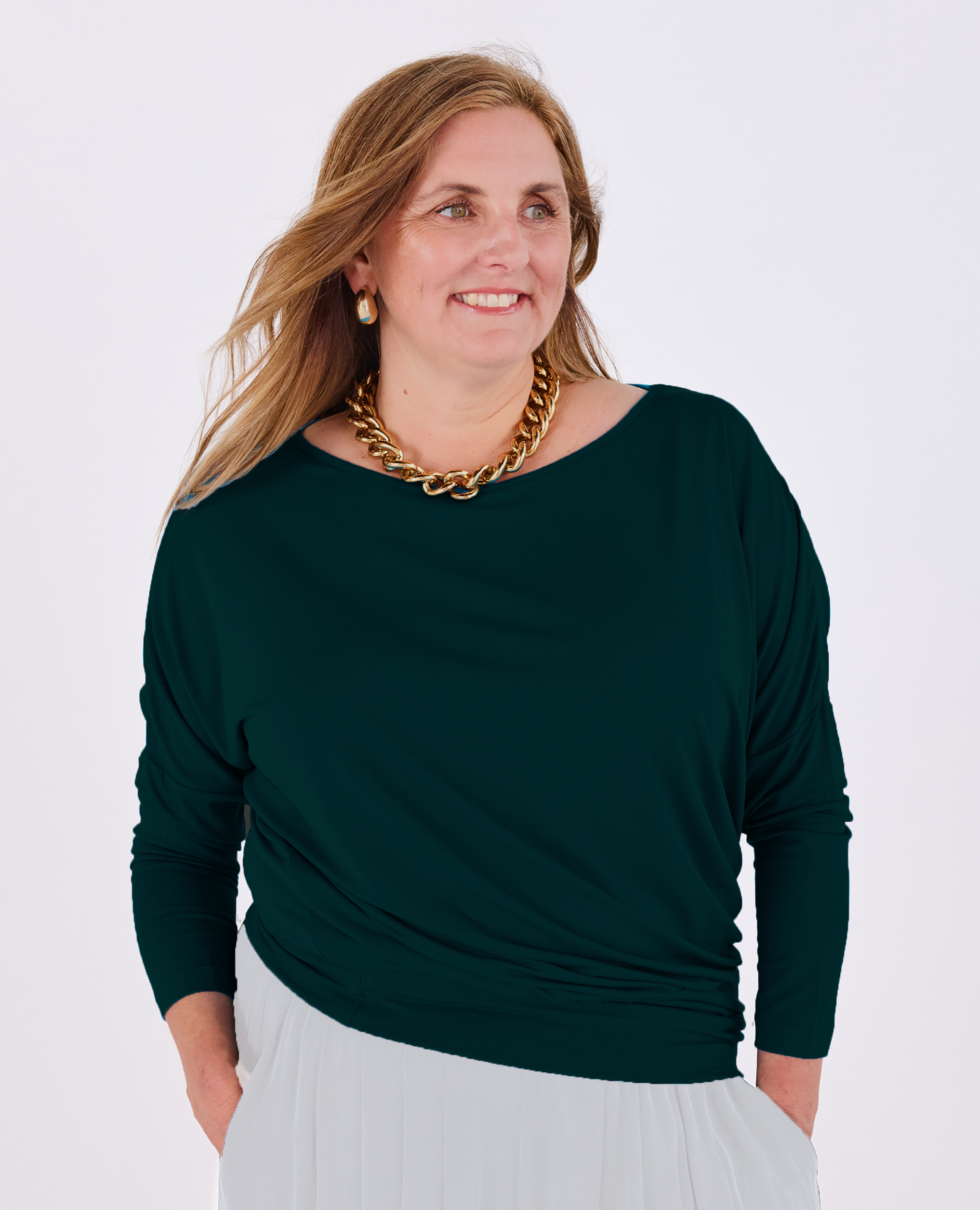 A woman wearing a dark green Melyr Aimee  long-sleeve boat-neck top and white pants, accessorized with a chunky gold chain necklace, photographed against a light grey studio background.