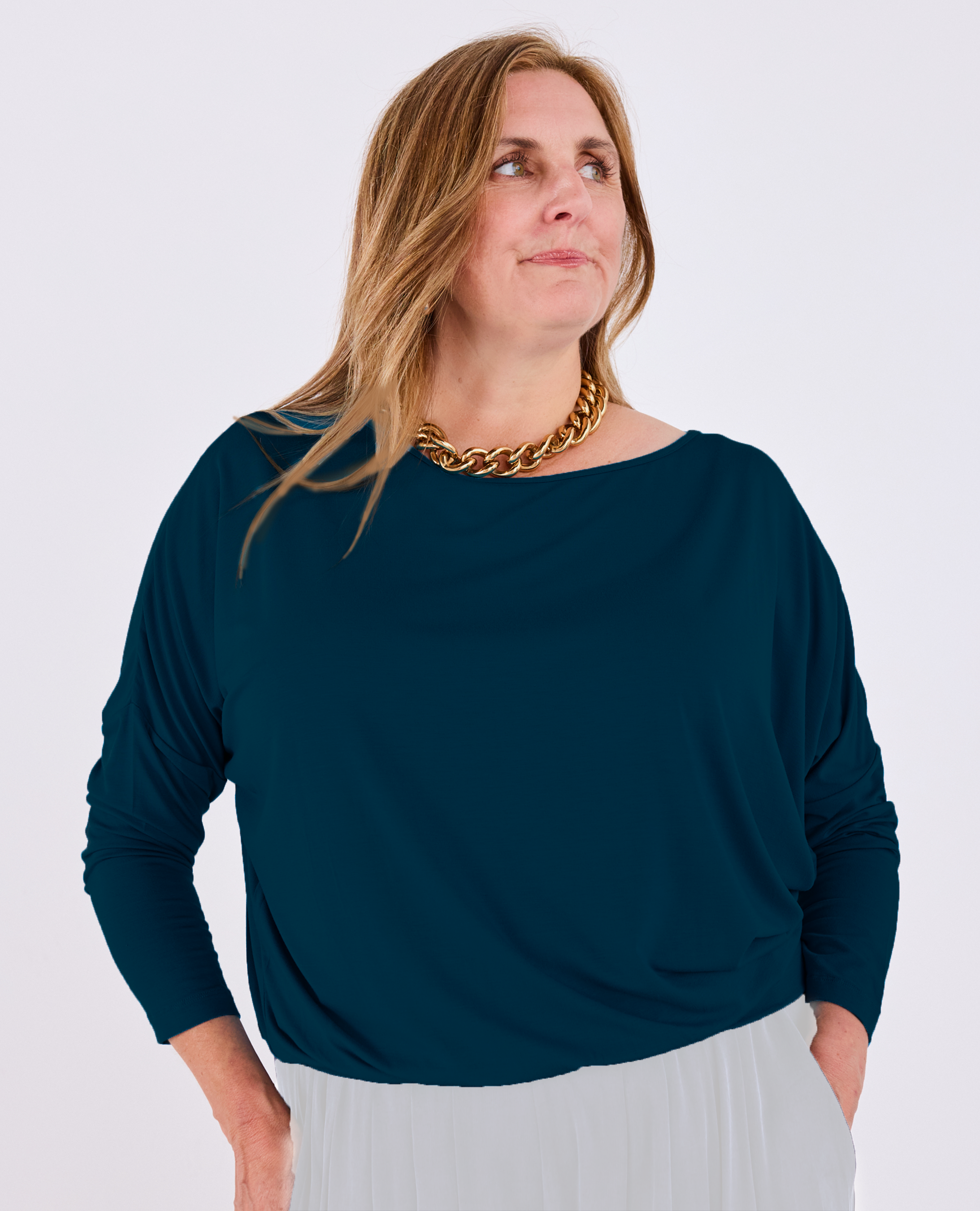 A woman wearing a dark teal long-sleeve Melyr Aimee batwing top and a chunky gold chain necklace, photographed against a light grey studio background.