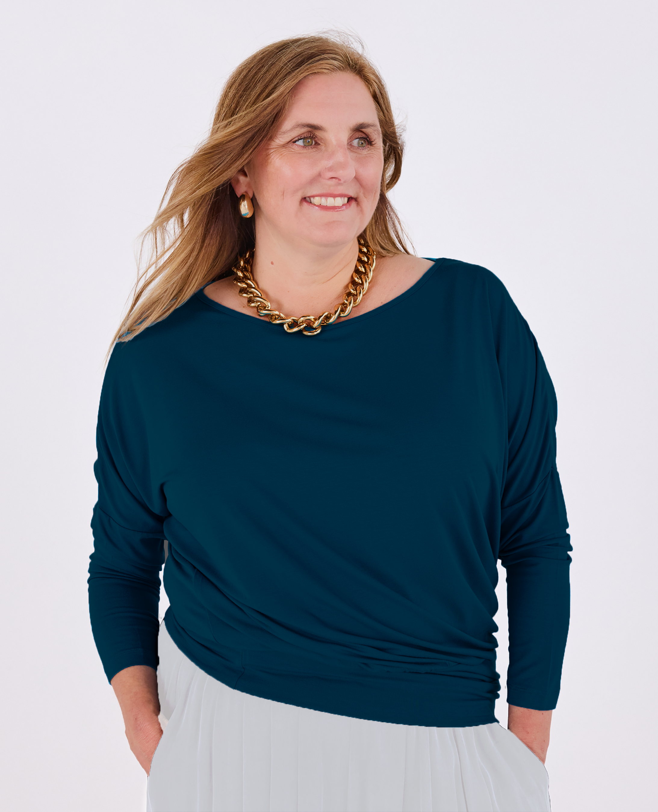 A woman wearing a dark teal long-sleeve Melyr Aimee batwing top and white skirt, accessorized with a chunky gold chain necklace, photographed against a light grey studio background.