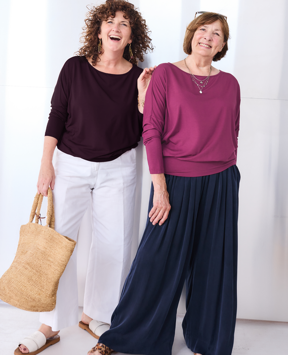 Two women modeling Melyr long-sleeve Aimee tops and wide-leg pants sets in a bright indoor setting. The woman on the left wears a dark plum Nico top with white pants and carries a woven straw tote bag.