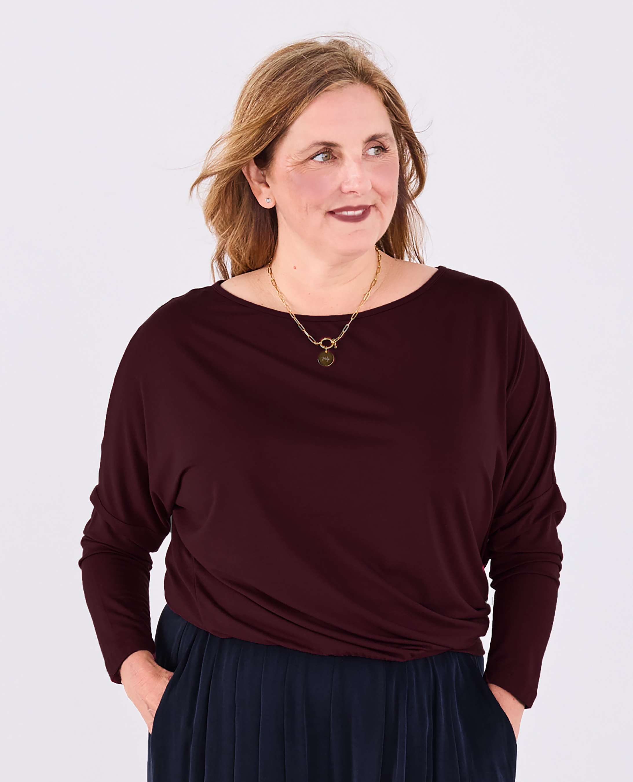 A model wearing a long-sleeve boat-neck batwing top by Melyr in deep burgundy, paired with a dark navy skirt, against a light grey studio background.