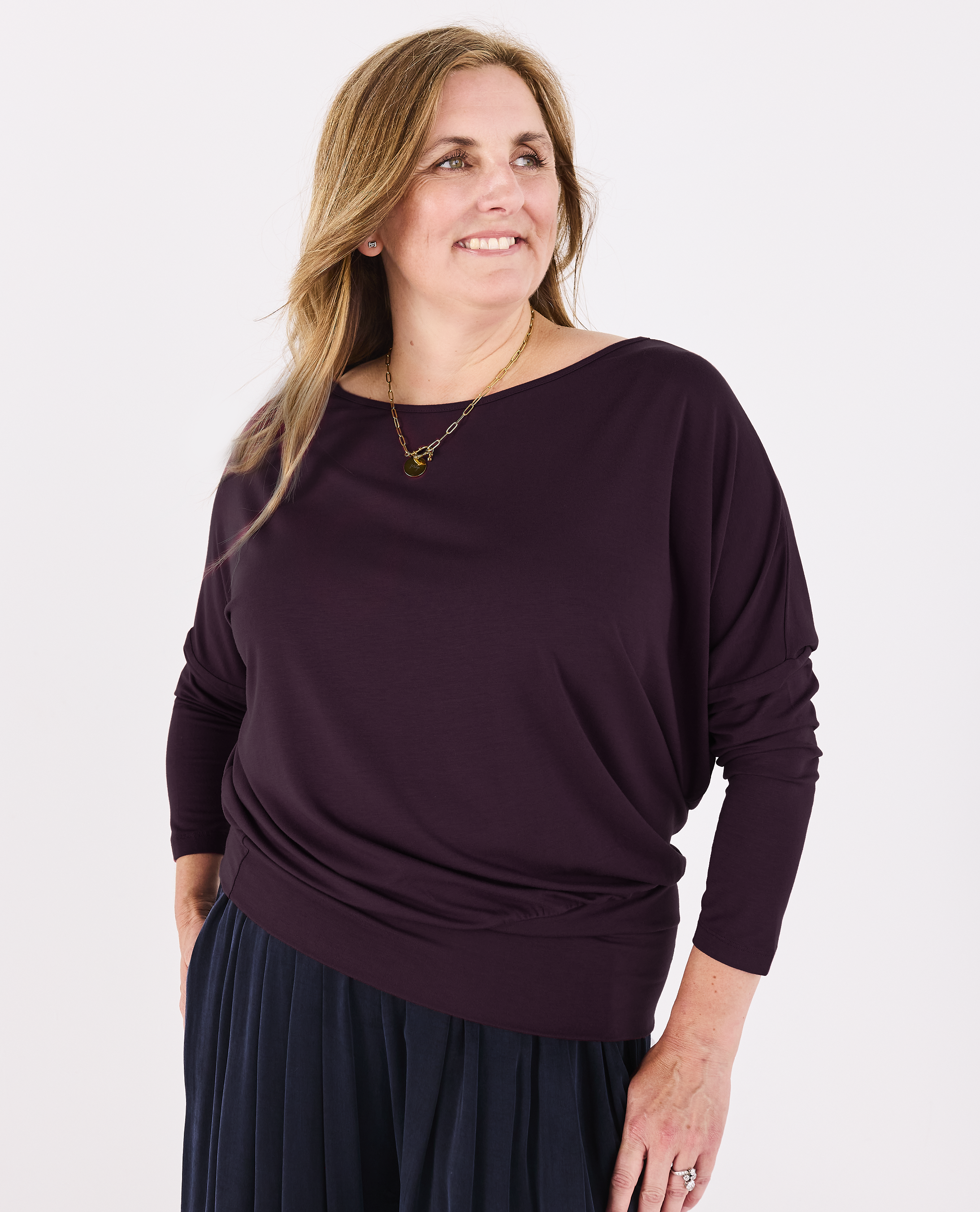 A woman wearing a dark plum Melyr Aimee long-sleeve batwing top and a dark navy pleated trousers, standing against a plain white background.