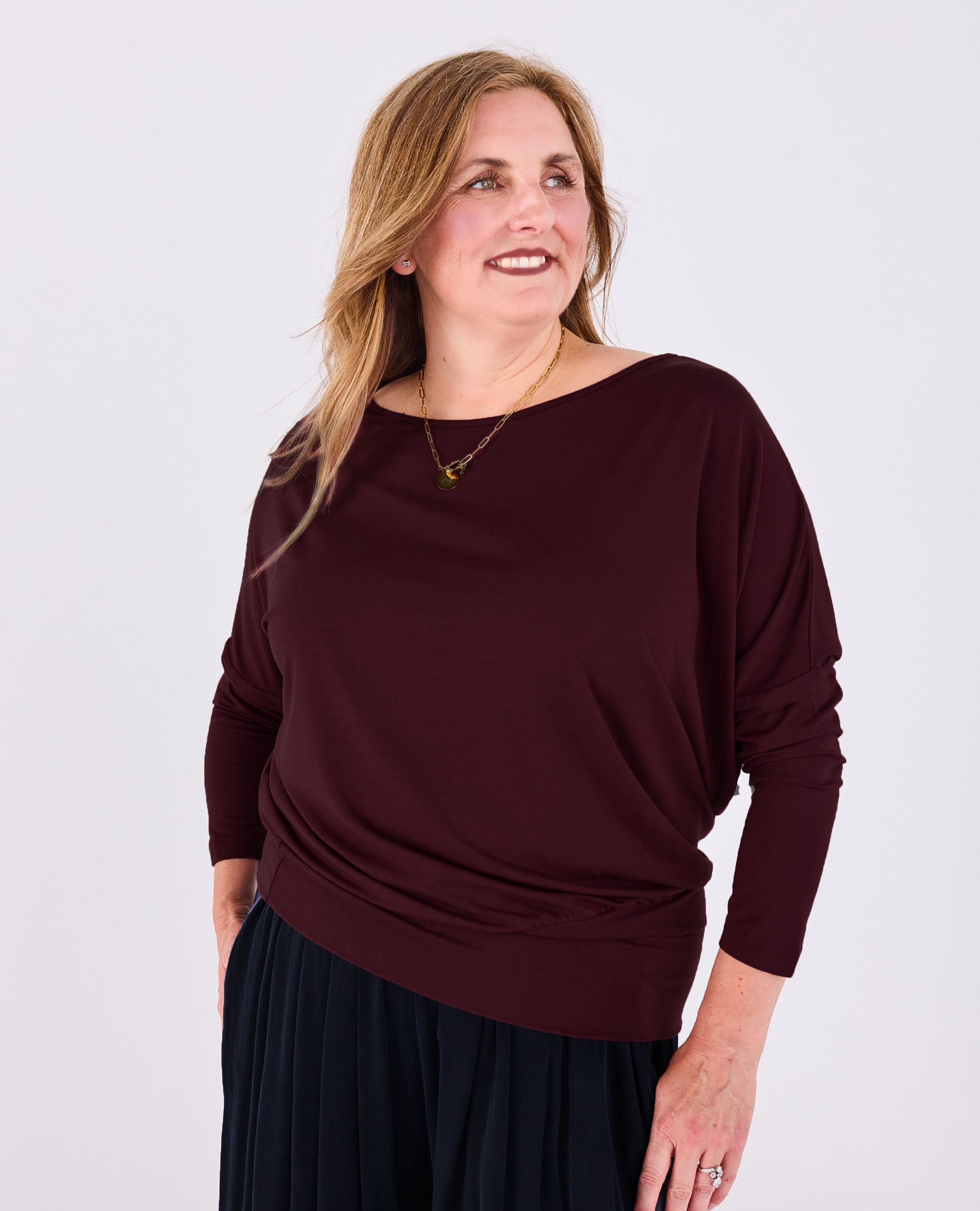 A woman wearing a dark burgundy long-sleeve Melyr batwing top and dark navy pleated skirt, standing against a light grey studio background.