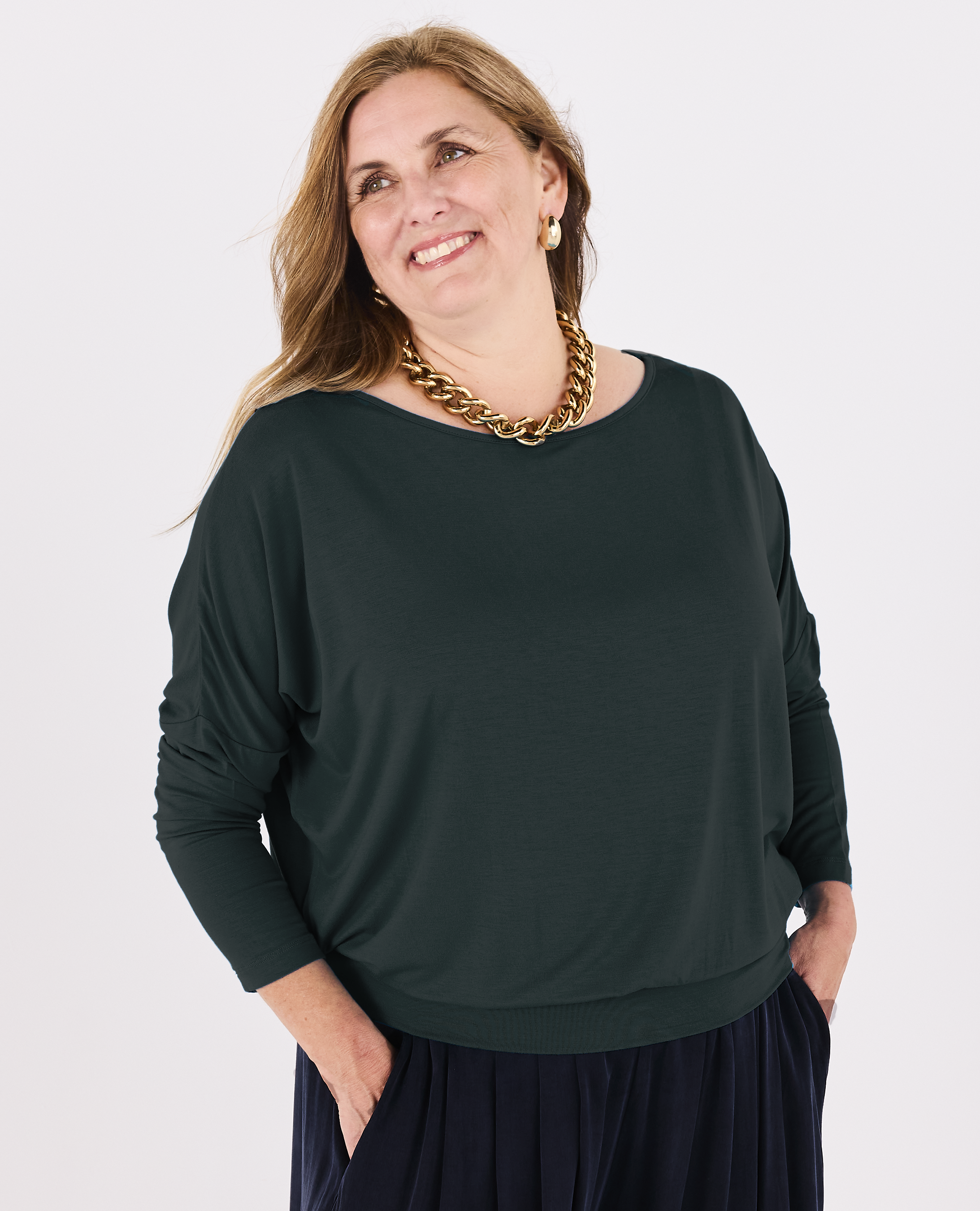 A woman wearing a Melyr Aimee dark green long-sleeve boat-neck top and a dark navy skirt, accessorised with a chunky gold chain necklace, against a plain white studio background.