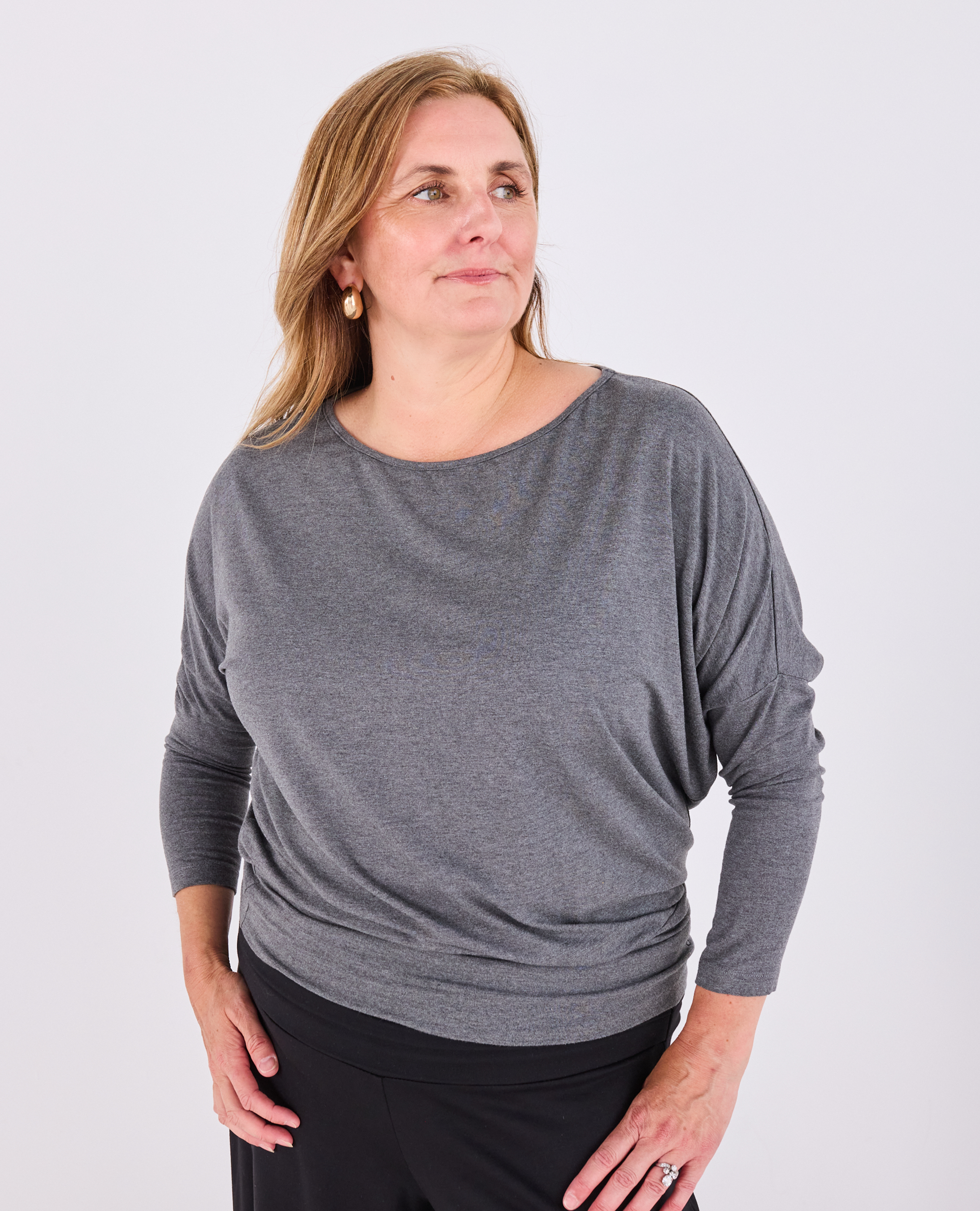 A woman wearing a heather gray long-sleeve boat-neck Melyr top and black pants, standing against a light gray studio background.