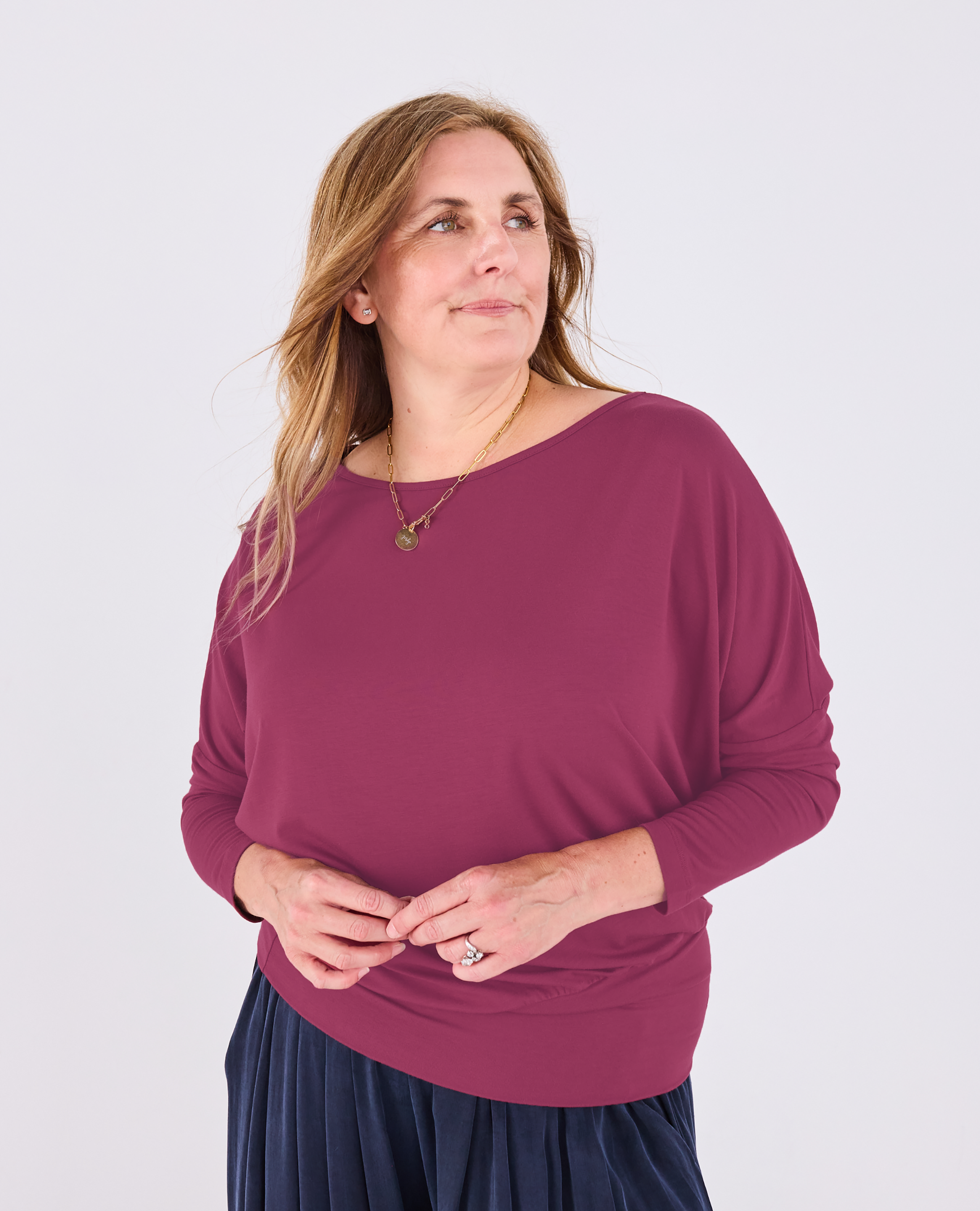A woman wearing a Melyr Aimee long-sleeve boat-neck top in a deep berry purple, paired with a dark navy pleated skirt, standing against a light gray studio background.