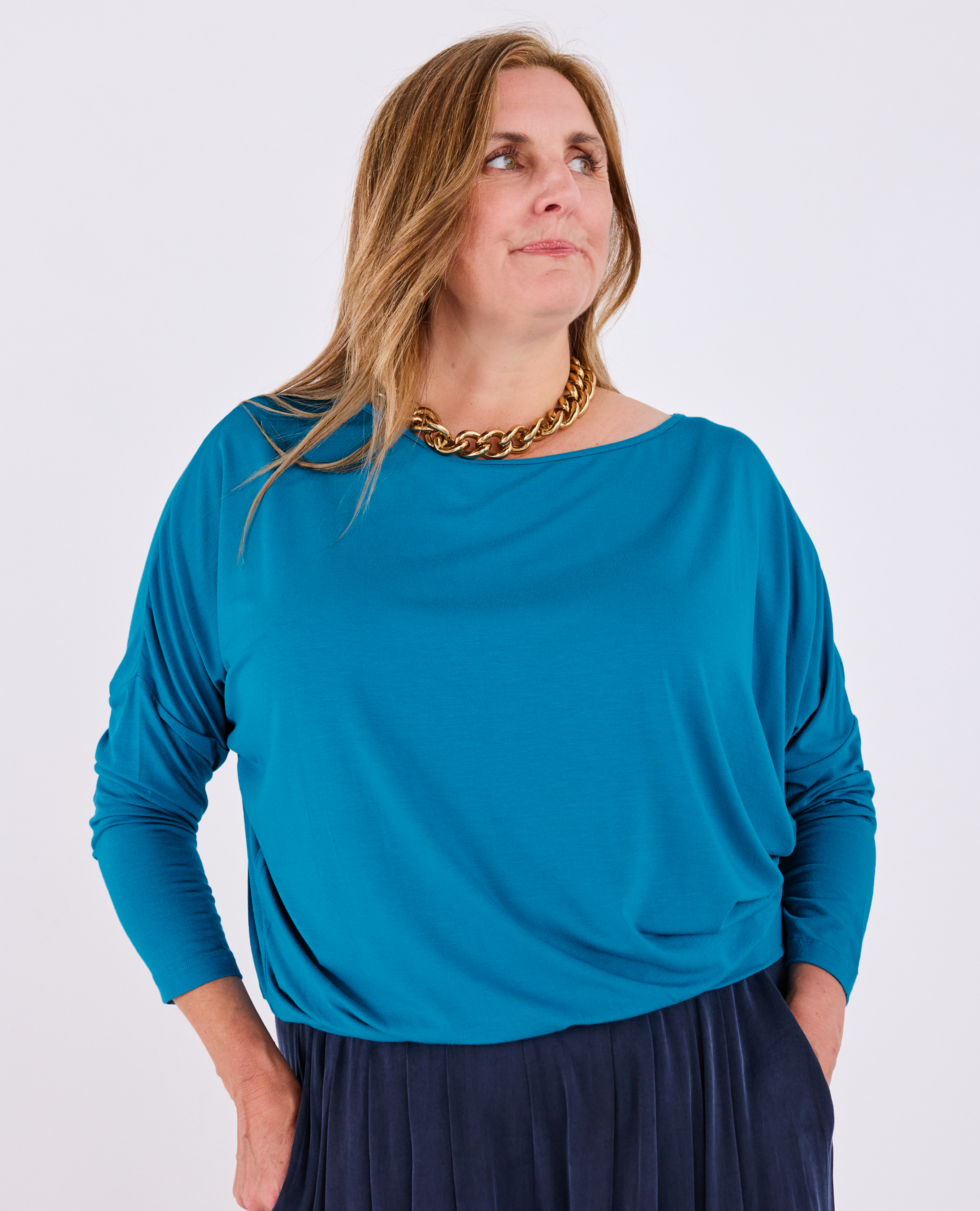 A woman wearing a bright teal blue long-sleeve Melyr Aimee batwing top and a dark navy pleated skirt, accessorized with a chunky gold chain necklace, photographed against a light grey studio background.