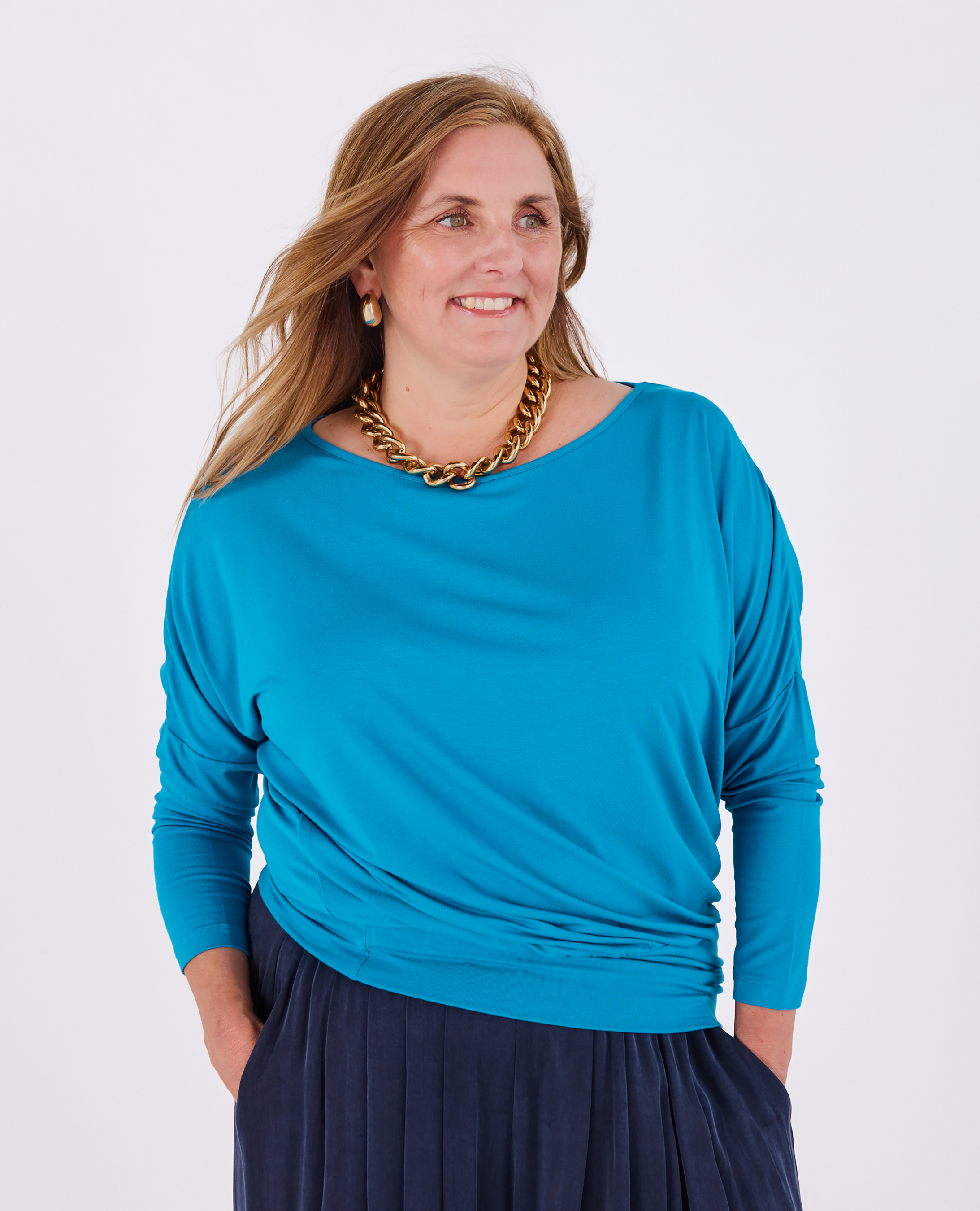 A woman wearing a teal blue long-sleeve top with a Melyr navy trousers accessorized with a chunky gold chain necklace, against a light gray background.