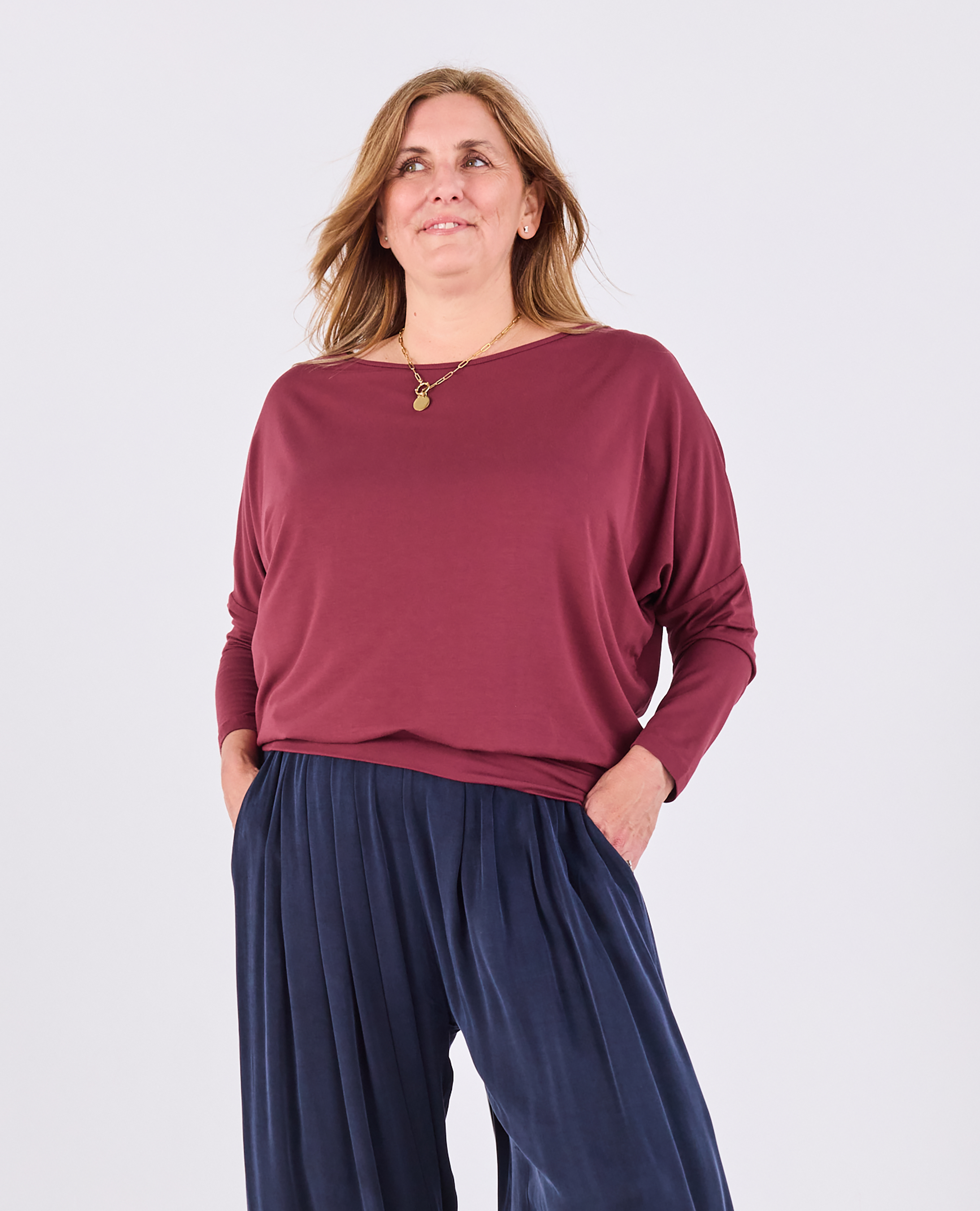 A woman modeling a long-sleeve boat-neck top in burgundy, paired with navy wide-leg pants, against a light gray studio background.
