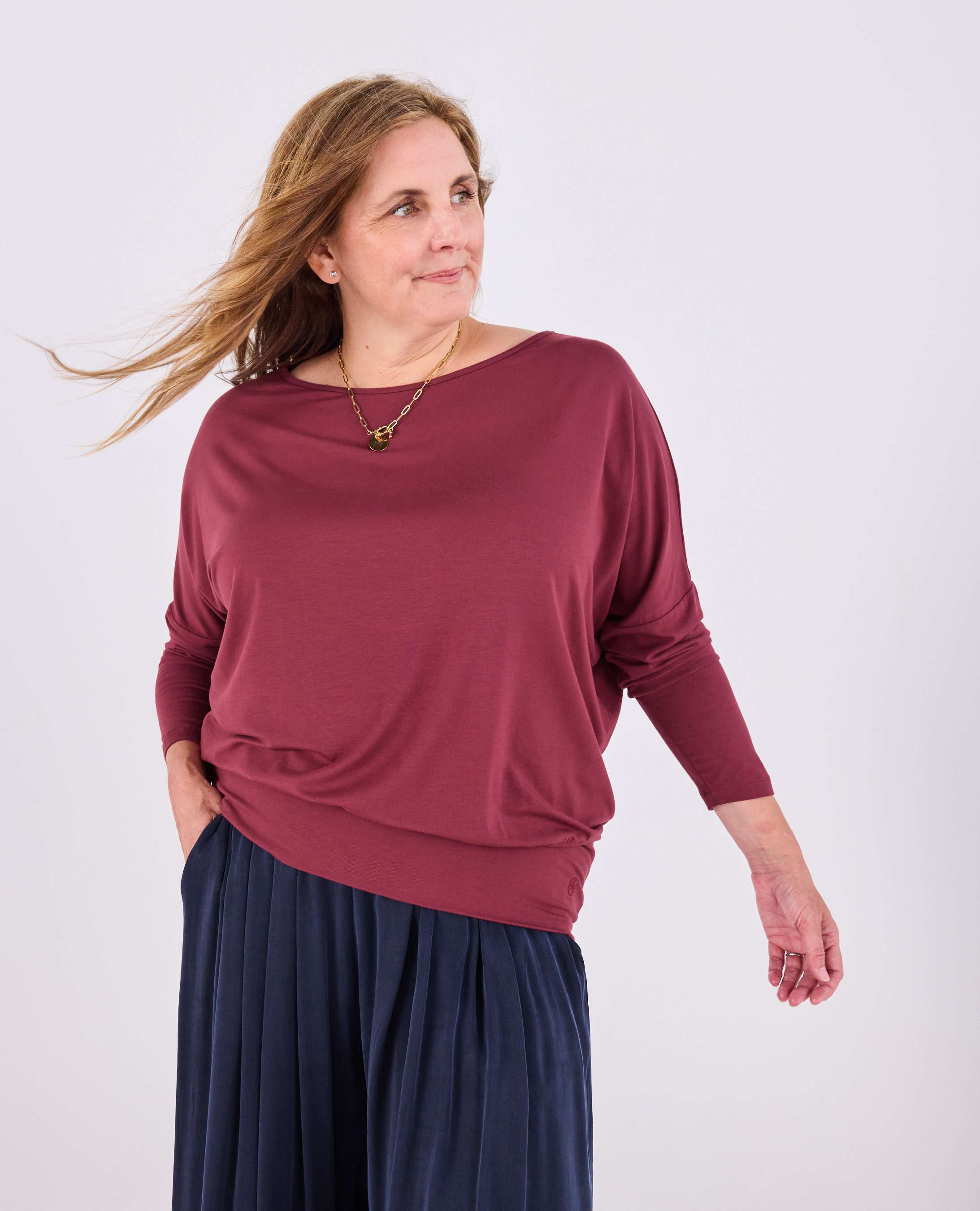 A woman modeling a long-sleeve boat-neck top in burgundy, paired with dark navy pleated pants, against a plain light gray studio background.