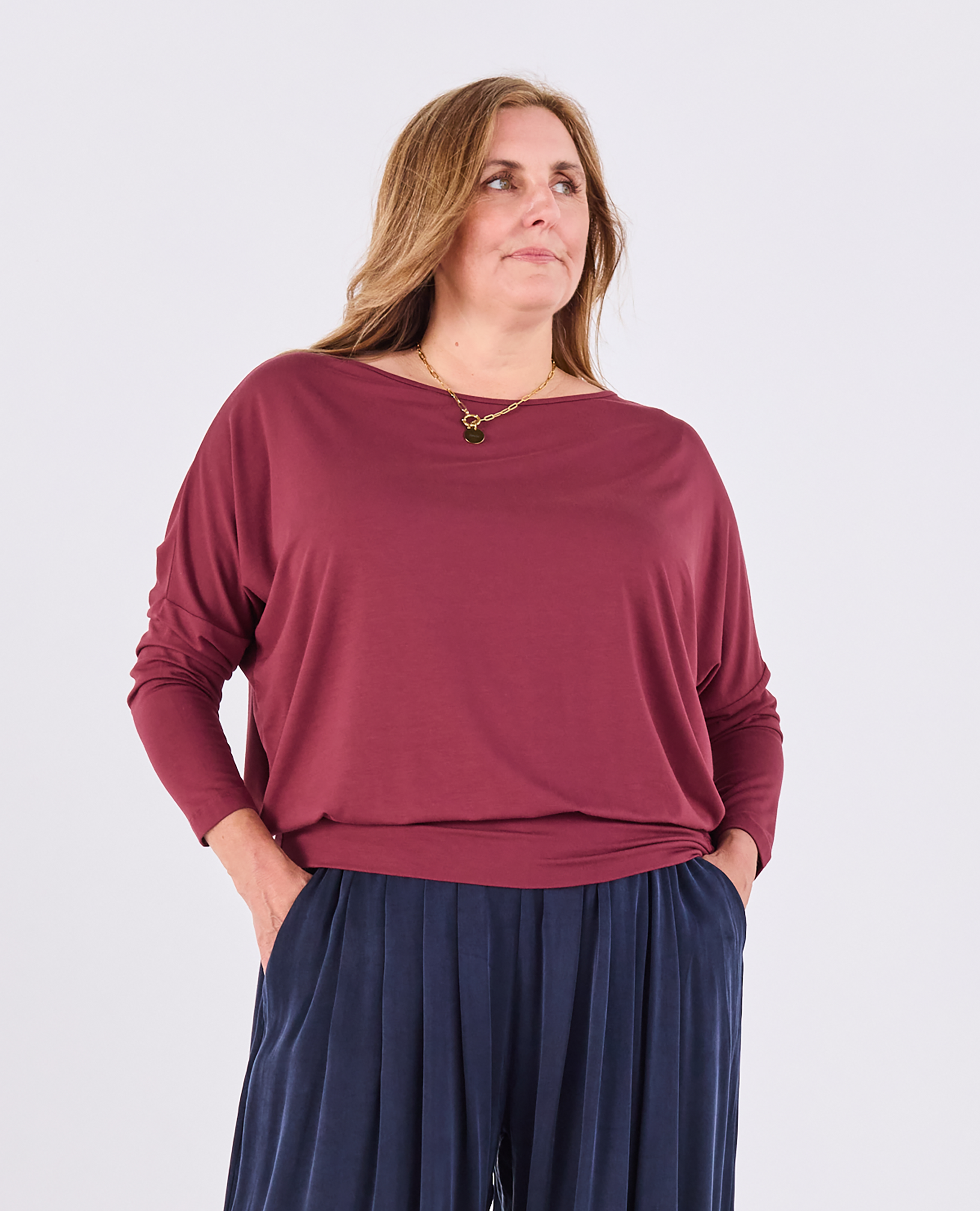 A woman wearing a loose-fitting, long-sleeve boat-neck top in a muted burgundy red, paired with dark navy pleated pants, against a light gray studio background.
