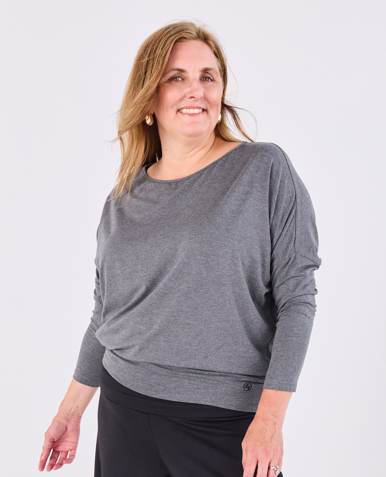 A woman wearing a heather gray long-sleeve boat-neck Melyr Aimee top and black pants, standing against a light gray studio background.