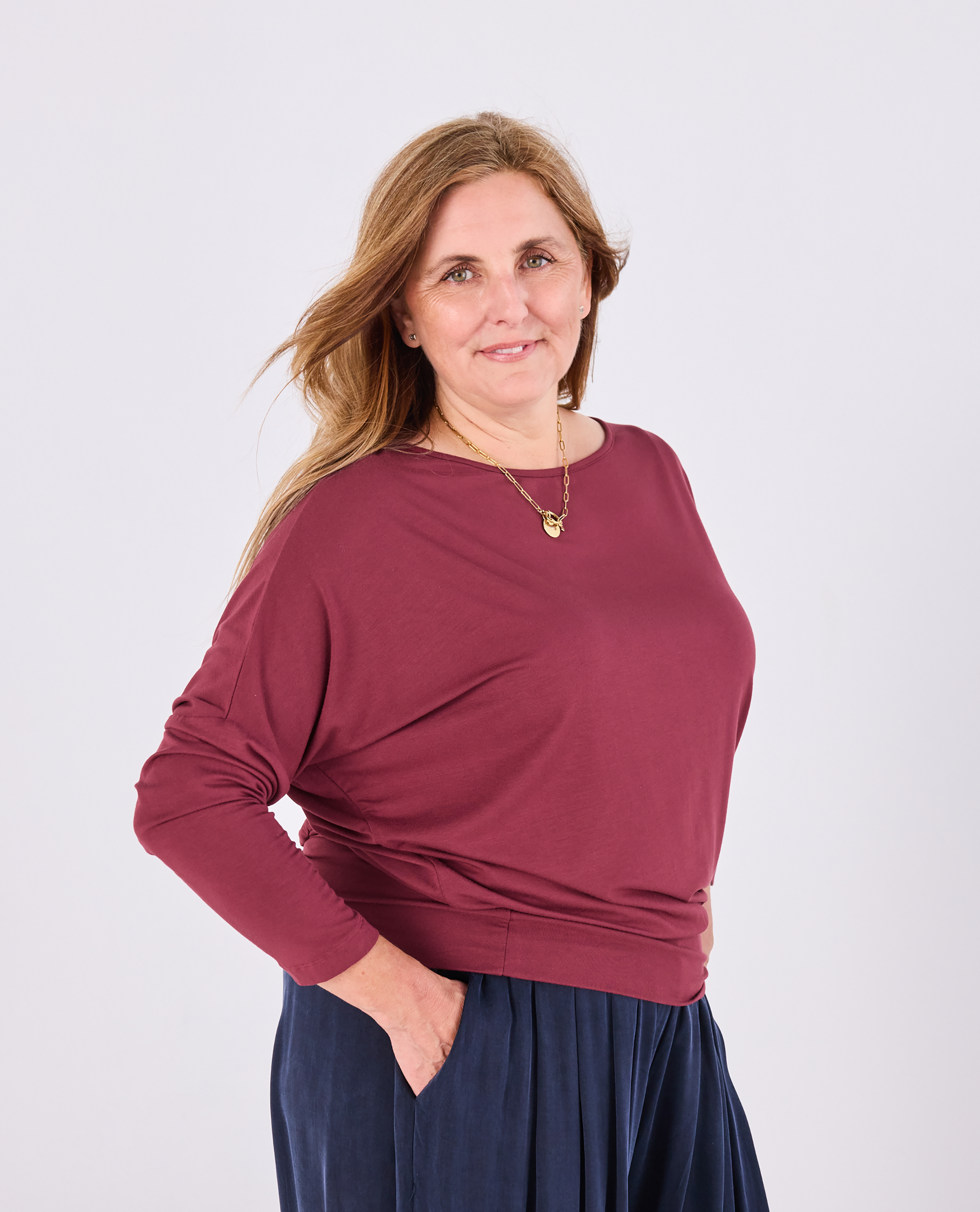 A woman modeling a long-sleeve boat-neck top in burgundy, paired with navy blue pants, against a plain light gray studio background.