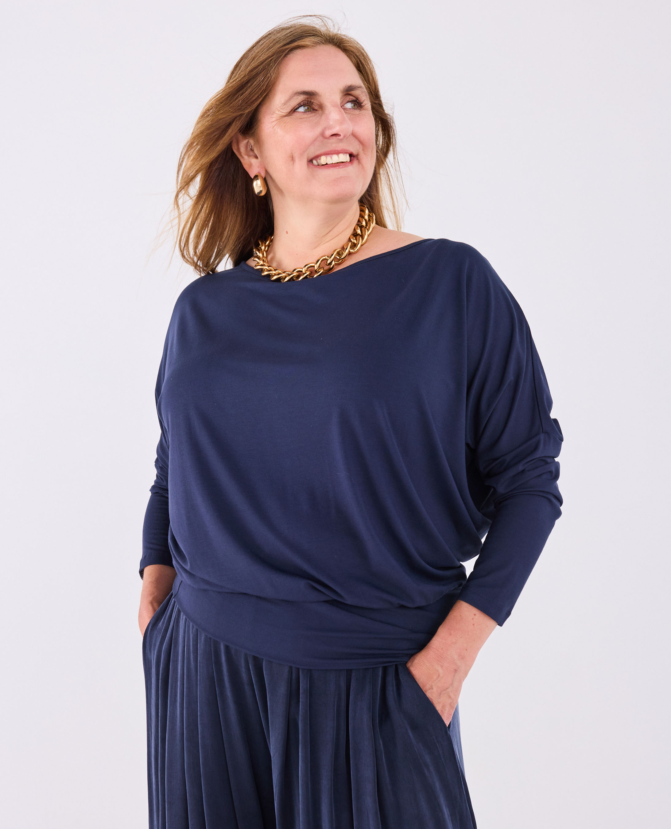 A woman wearing a Melyr navy Aimee long-sleeve draped top and matching pleated skirt, accessorized with a chunky gold chain necklace, against a light grey studio background.