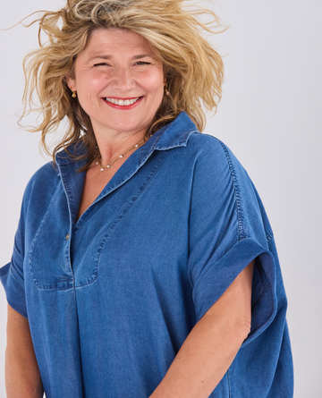 A smiling blonde woman wearing a Melyr Alessia, short-sleeve blue denim-style button-up shirt against a plain light gray studio background.