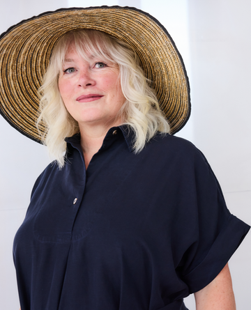 A woman with blonde hair wearing a large woven straw sun hat and a Melyr dark navy blue short-sleeve button-up shirt, photographed against a plain white background.