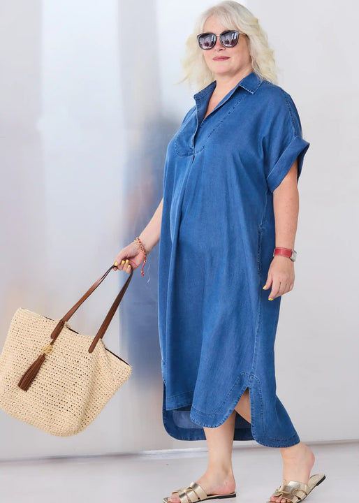 A woman modeling a Melyr Alessia blue denim shirt dress, holding a large woven straw tote bag with brown handles against a light gray studio background.