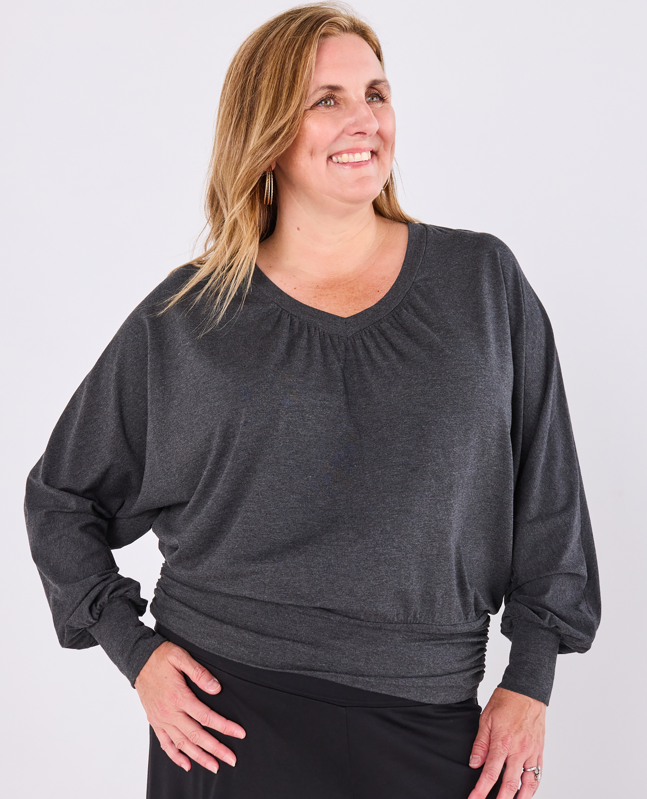 A plus size woman wearing a dark grey Melyr vivienne long-sleeve top with a gathered v-neckline and ruched hem, paired with black bottoms, photographed against a light grey studio background.