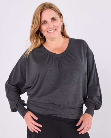 A smiling blonde woman wearing a dark charcoal grey long-sleeve Melyr Vivienne top with a gathered v-neckline and black pants, standing against a light grey studio background.
