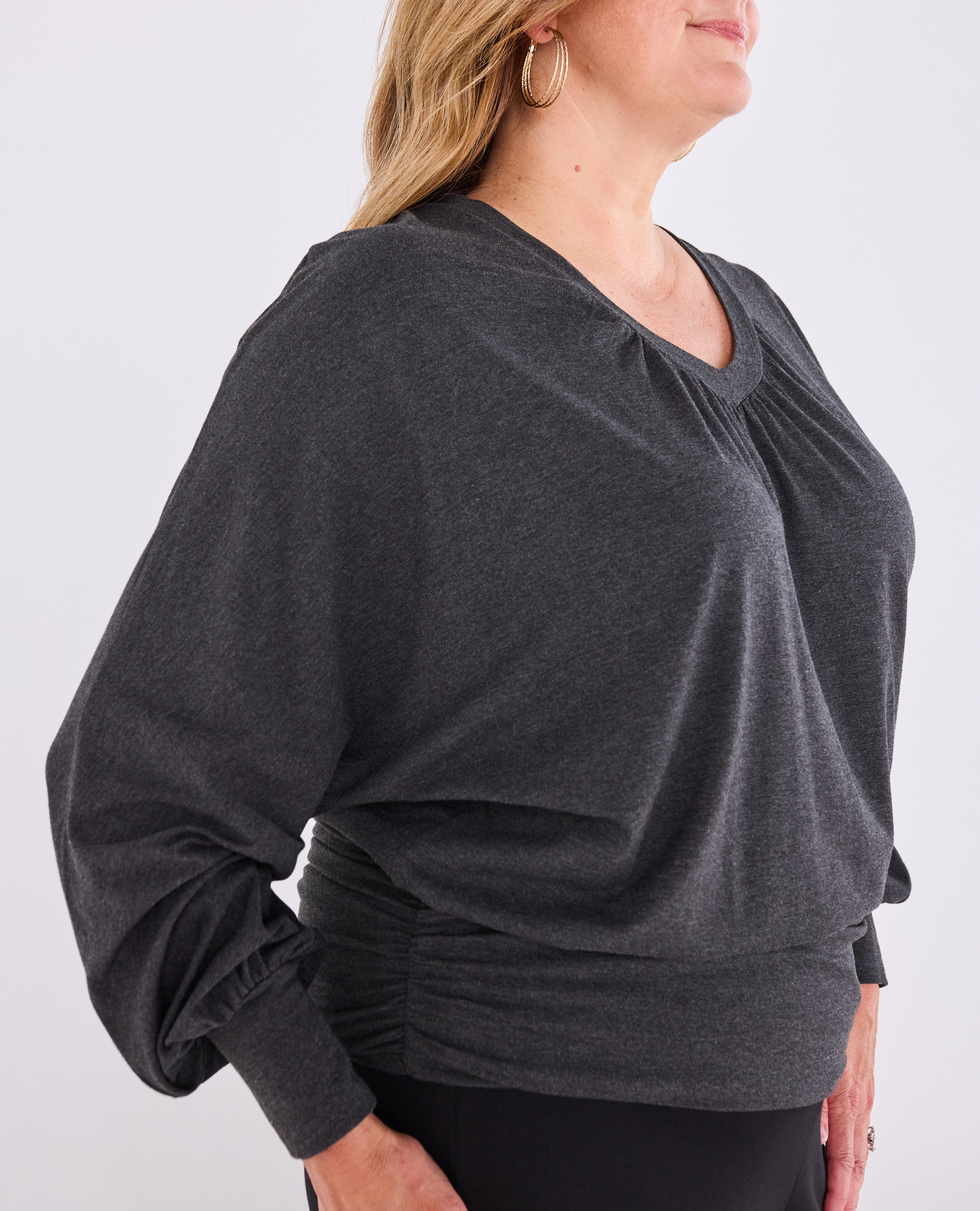 A woman wearing a dark charcoal gray long-sleeve Melyr Vivienne top with a gathered, ruched neckline and a matching ruched waist detail, paired with black bottoms against a light grey studio background.
