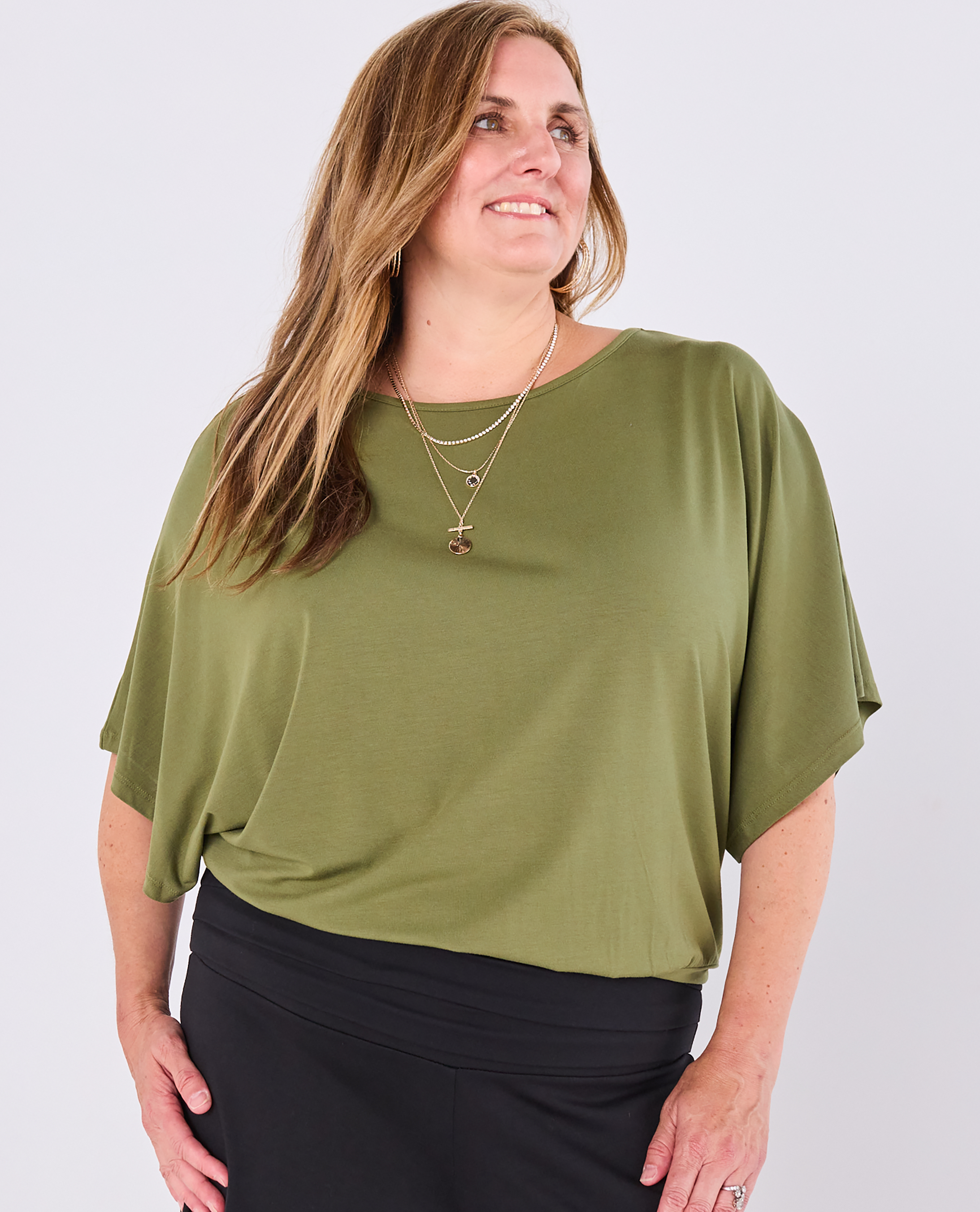 A woman wearing a Melyr olive green short-sleeve Nico top and black bottoms, standing against a light grey studio background.