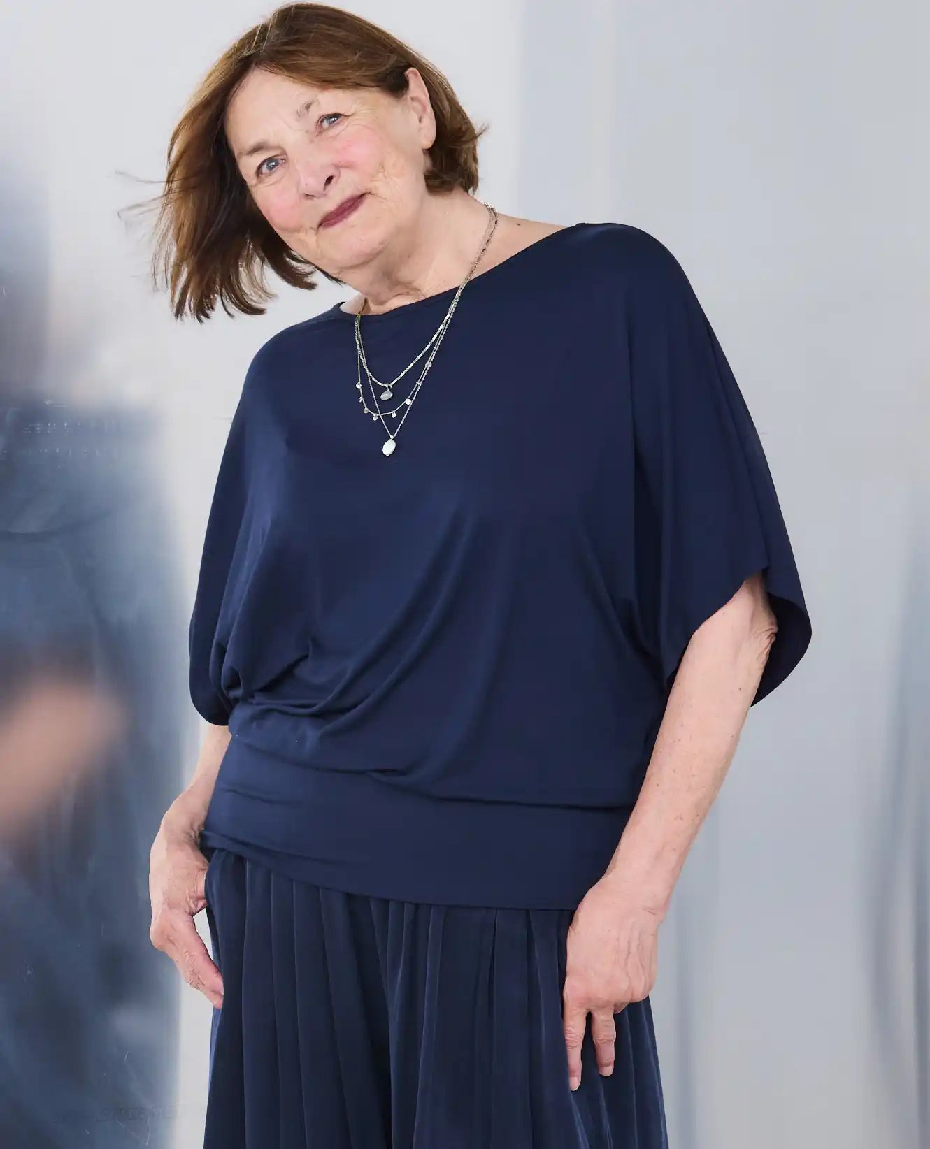 A woman wearing a Melyr navy Nico draped short-sleeve top and matching pleated skirt, accessorized with layered silver necklaces, photographed against a soft silver background.