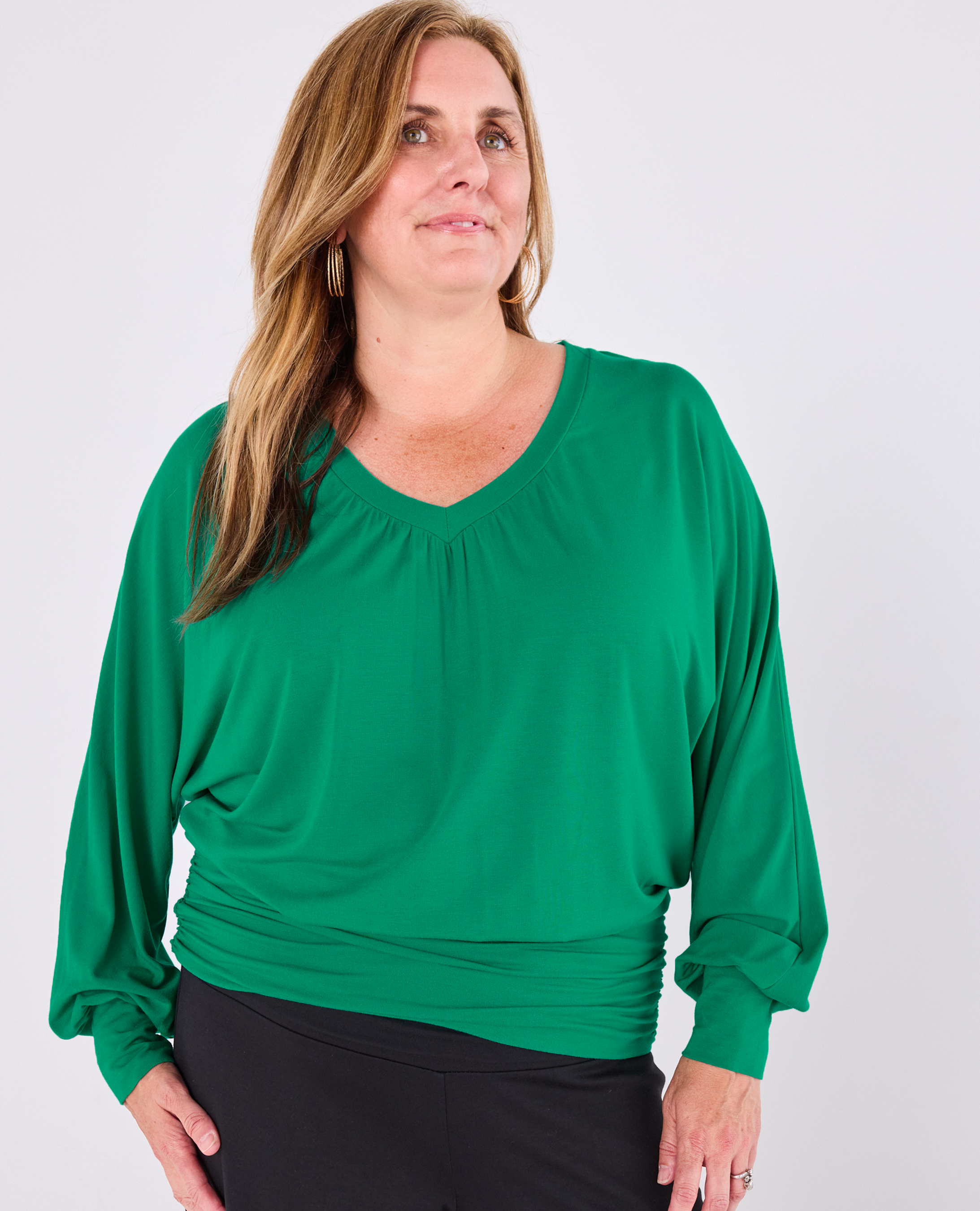 A woman wearing a bright green Melyr vivienne long-sleeve top with a gathered V-neck and ruched hem, paired with black pants, against a light gray studio background.