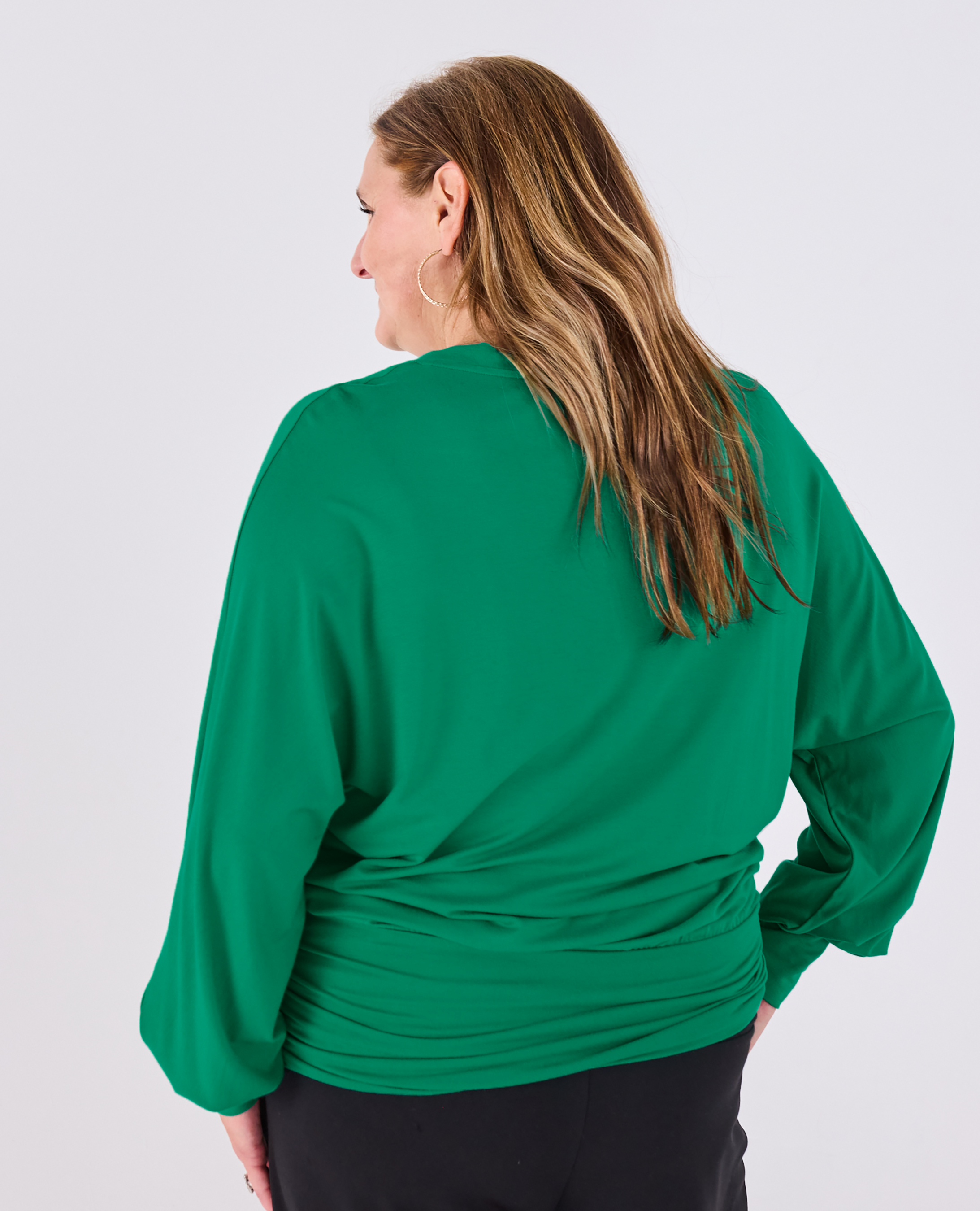 A woman shown from the back wearing a Melyr bright green Vivienne long-sleeve top and black pants against a light gray studio background.