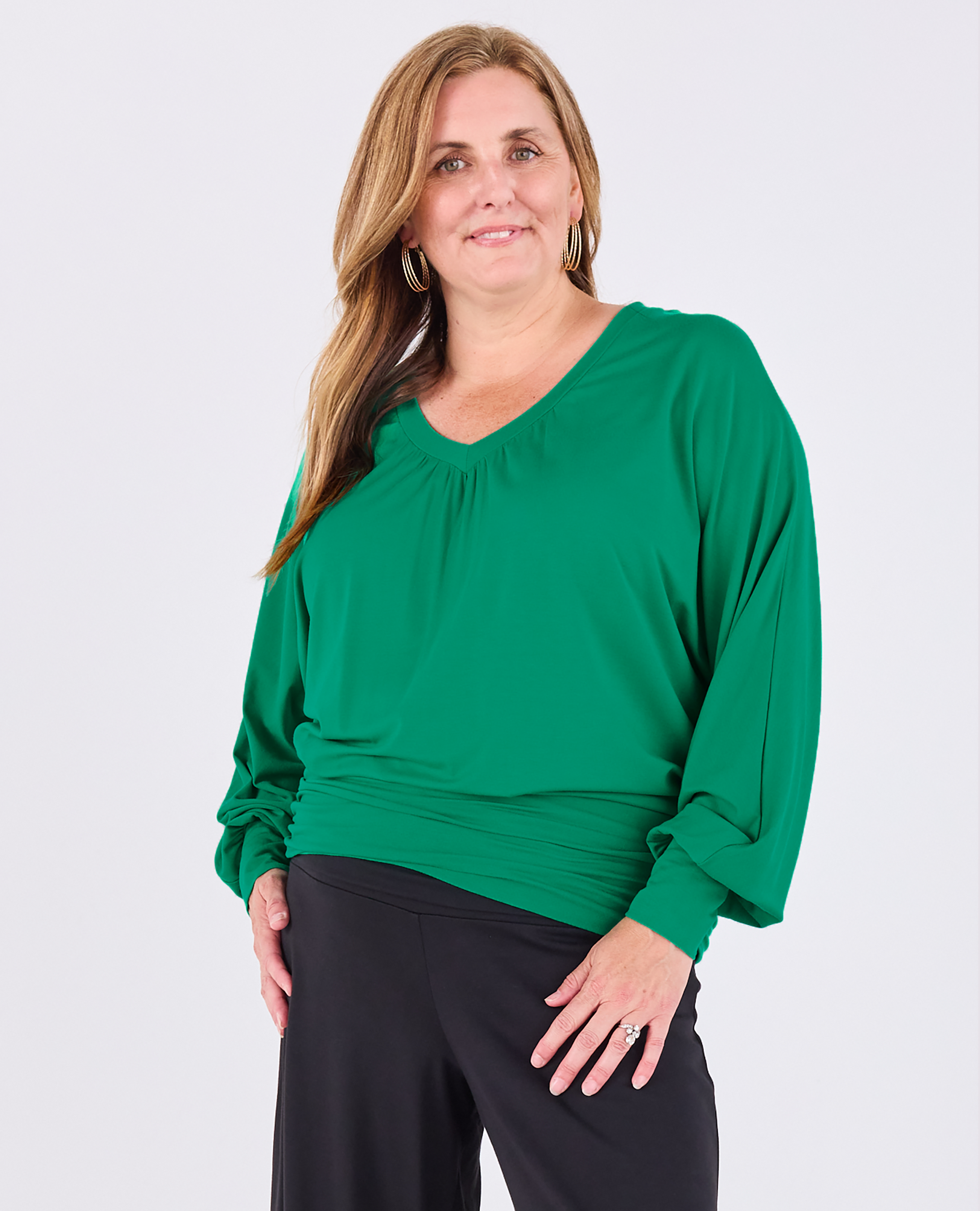 A woman modeling a Melyr vibrant green Vivienne long-sleeve top with a gathered neckline and draped sleeves, paired with black pants, against a plain light gray background.