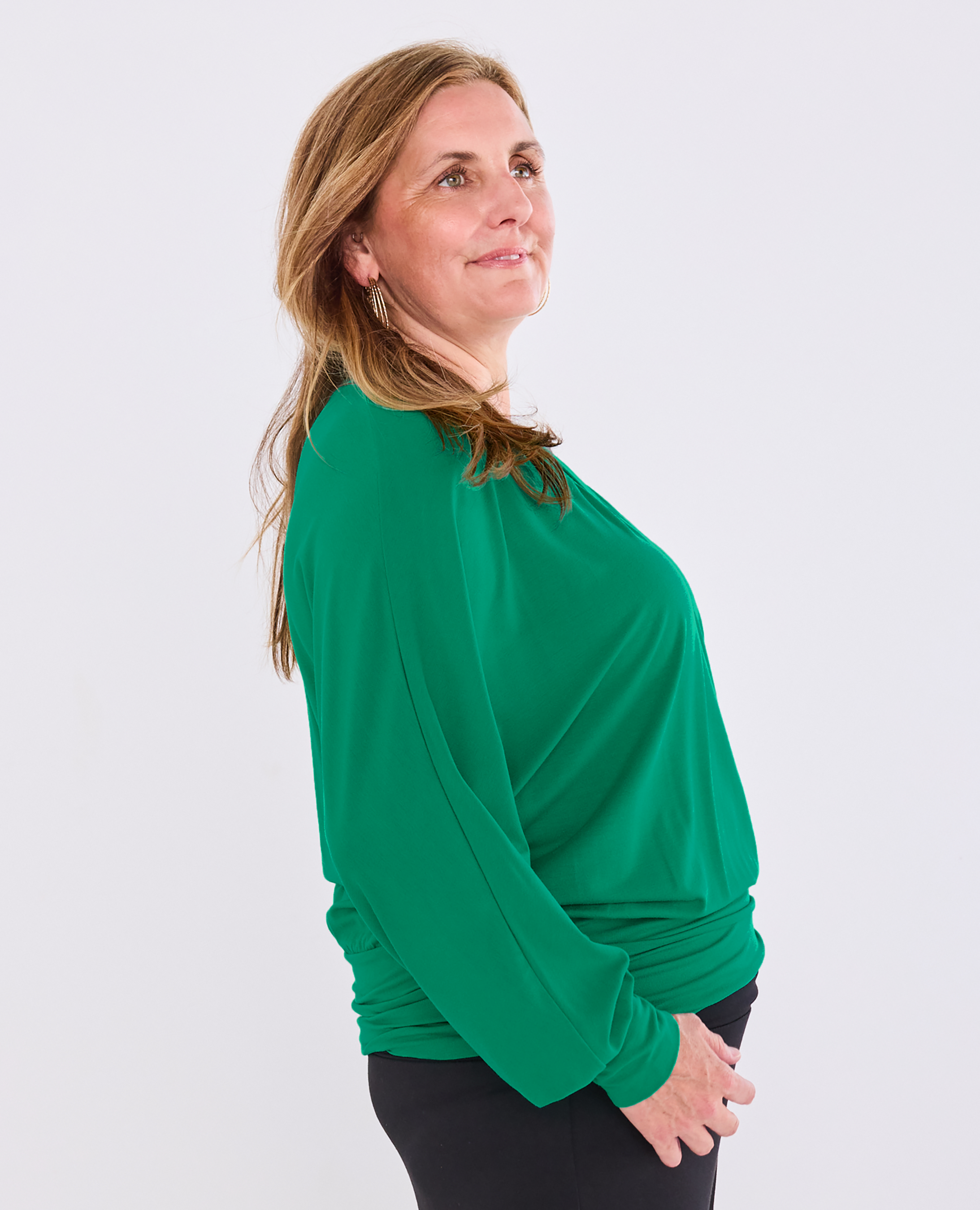 A woman wearing a Melyr bright green long-sleeve top and black pants, shown from a side profile against a light gray studio background.