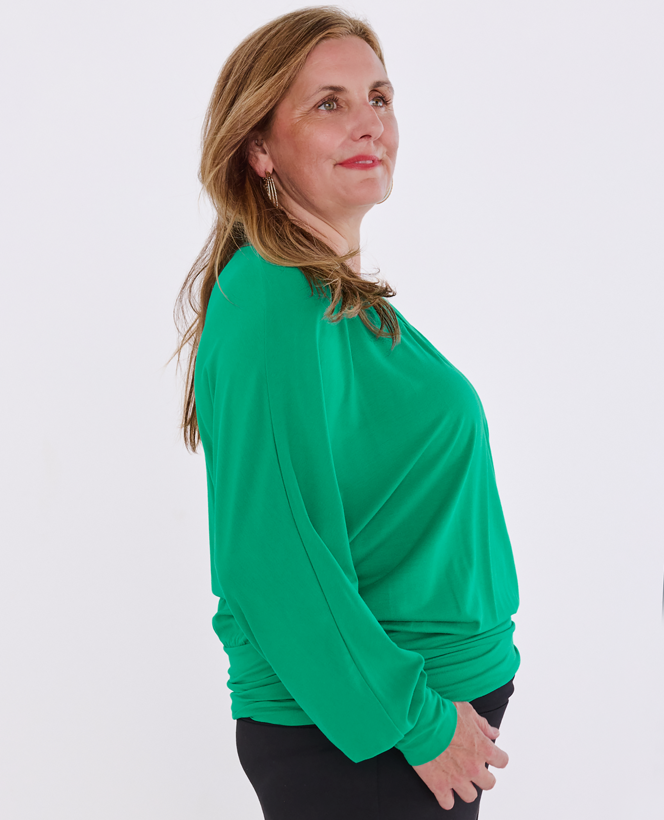 A woman wearing an emerald green Melyr Vivienne batwing long-sleeve top with a gathered, ruched hem, shown in profile against a plain light grey background.