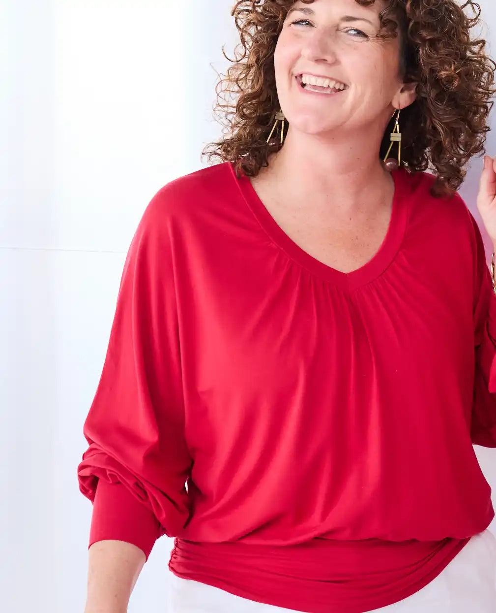A woman wearing a bright red, Melyr Vivienne long-sleeve V-neck blouse with a gathered, pleated chest design and a slightly draped silhouette. She is smiling and looking off-camera against a clean white background.