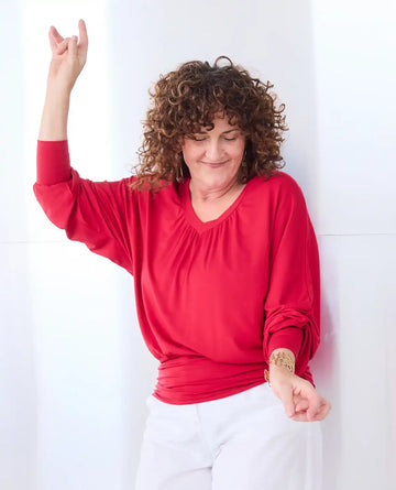A woman with curly brown hair wearing a bright red Melyr vivienne long-sleeve top and white pants, posing with one arm raised against a white wall.
