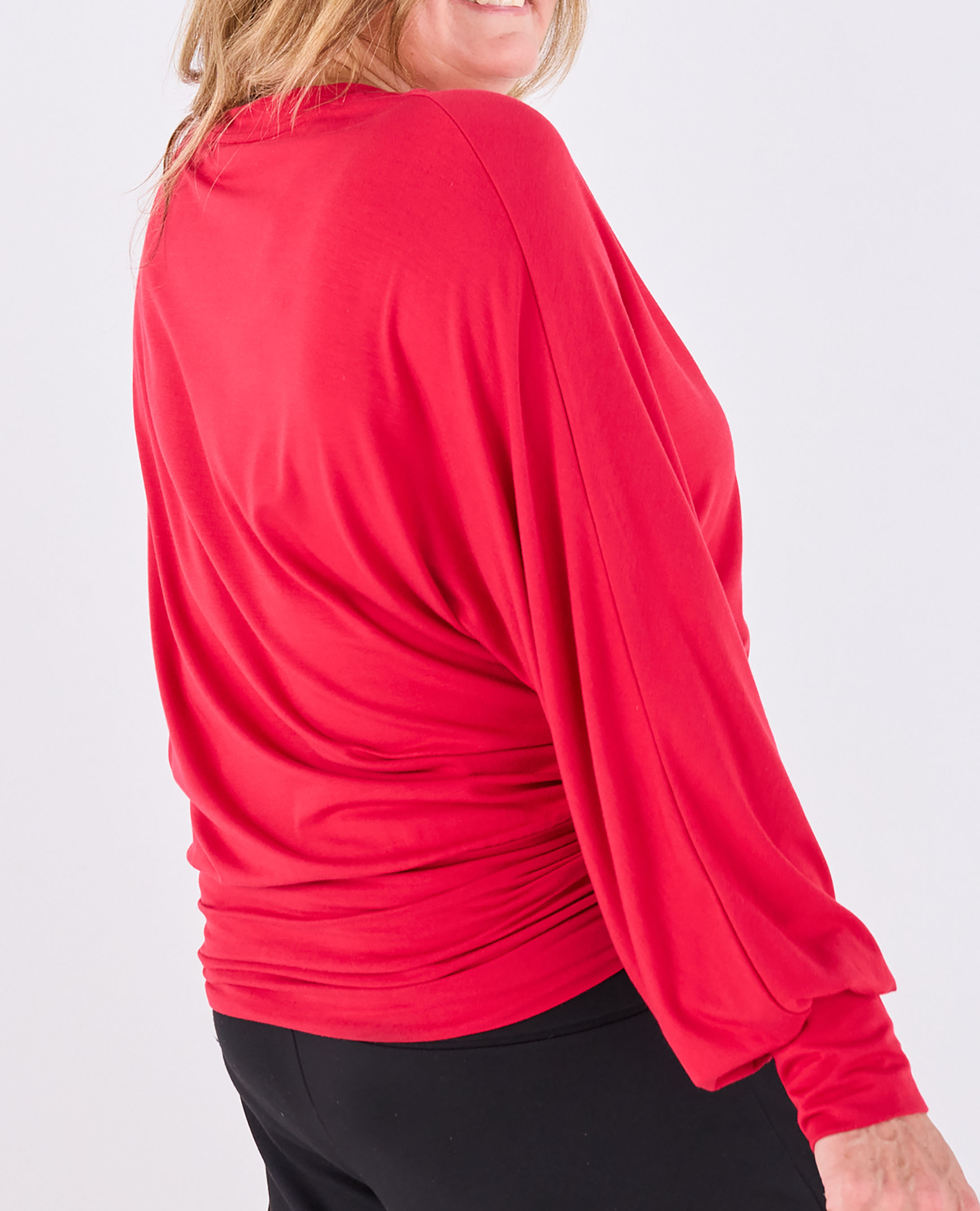 A woman wearing a bright red long-sleeve Melyr Vivienne top with a draped, elbow-length sleeve and gathered side detail, paired with black bottoms, shown from a side-back angle against a light gray studio background.