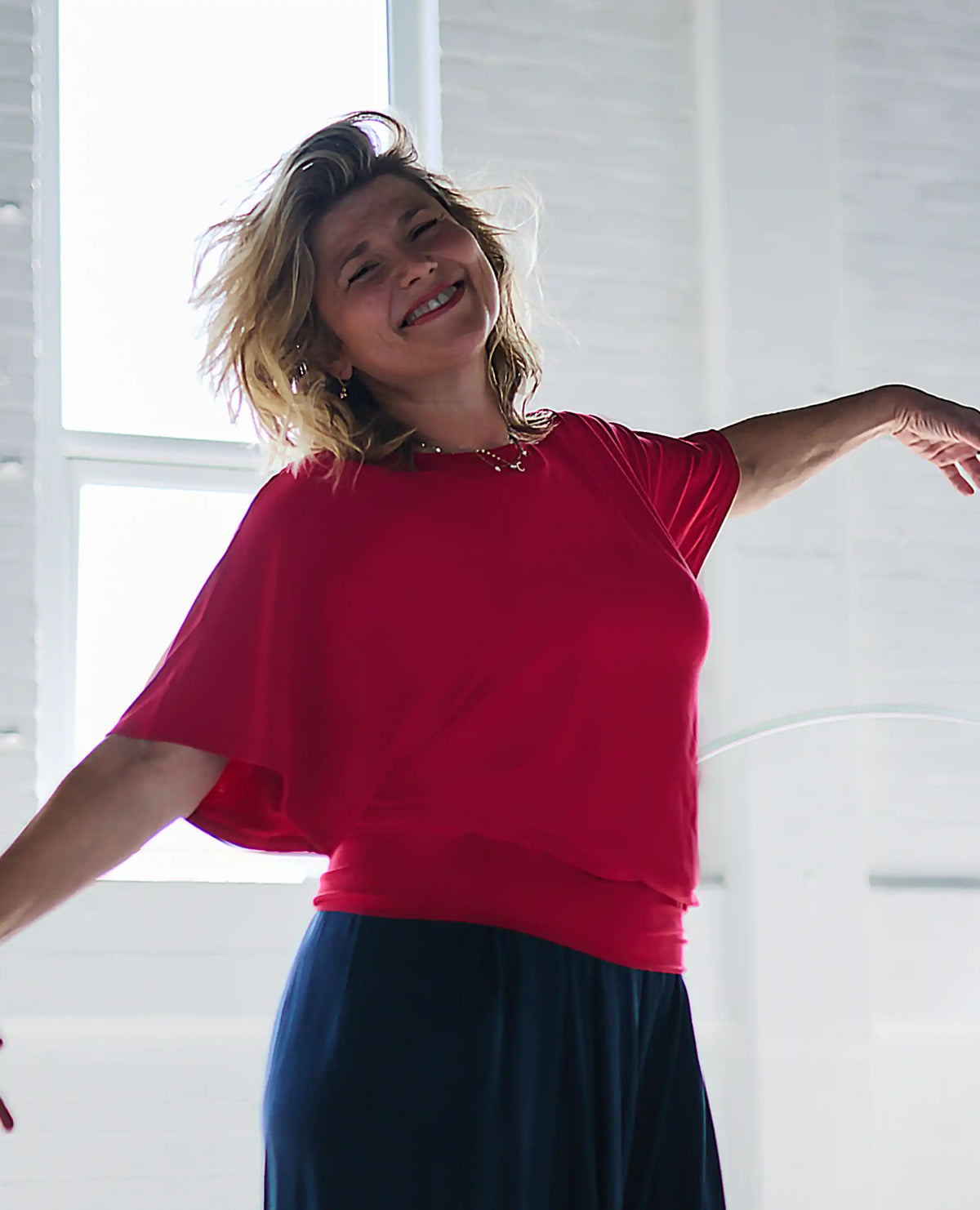 A woman with blonde hair smiling and looking upward, wearing a Melyr bright red short-sleeve top and a dark navy skirt, posed in a bright white room with window light.
