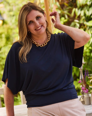 Woman wearing a Melyr Nico navy blue top and Melyr Amalfi Camel pants with a gold necklace, standing outdoors.