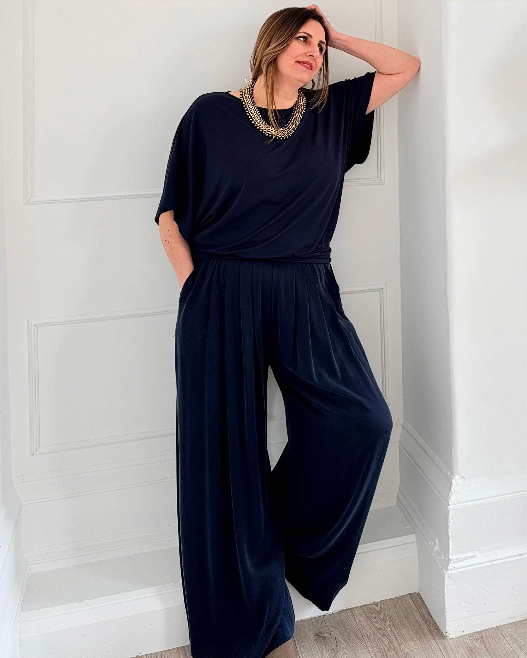 Woman wearing a navy blue trousers and top from Melyr leaning against a white paneled wall.