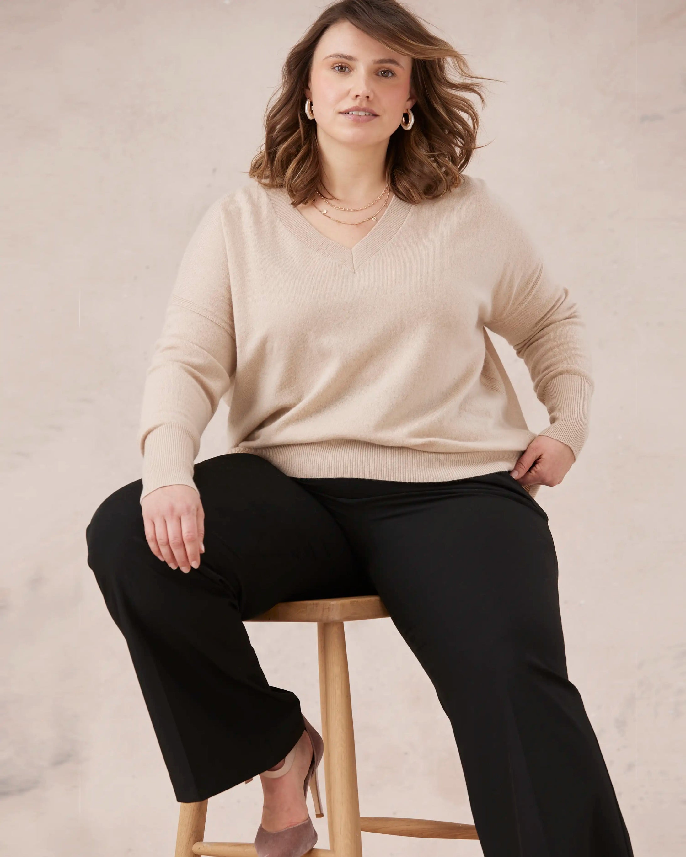 Woman sitting on a stool wearing a beige melyr knitted sweater and black pants against a neutral background